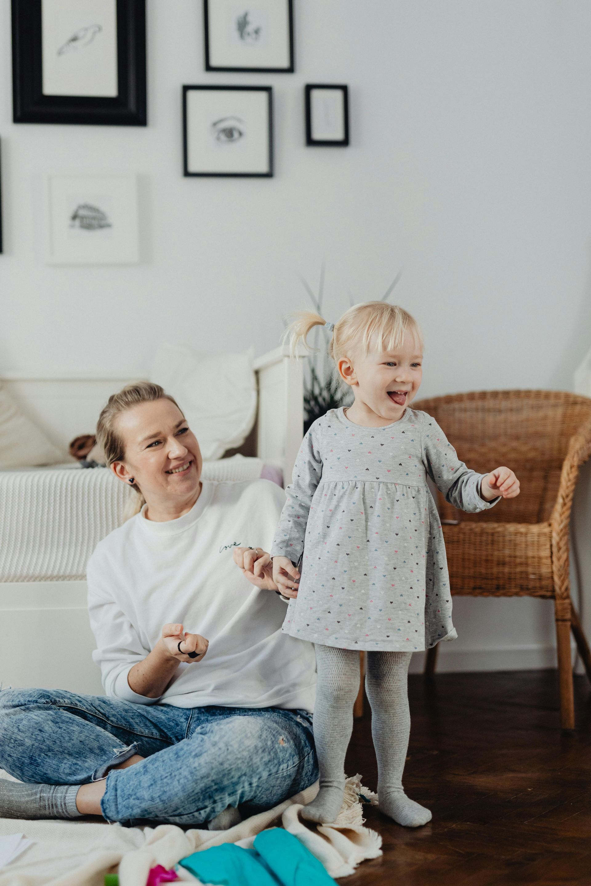 Person and child in a bright living room, sitting and standing near a wicker chair and framed wall art.