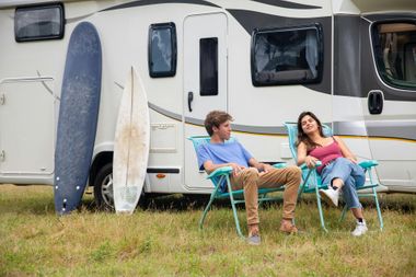 Two people relaxing in chairs beside a camper, with surfboards leaning against it.