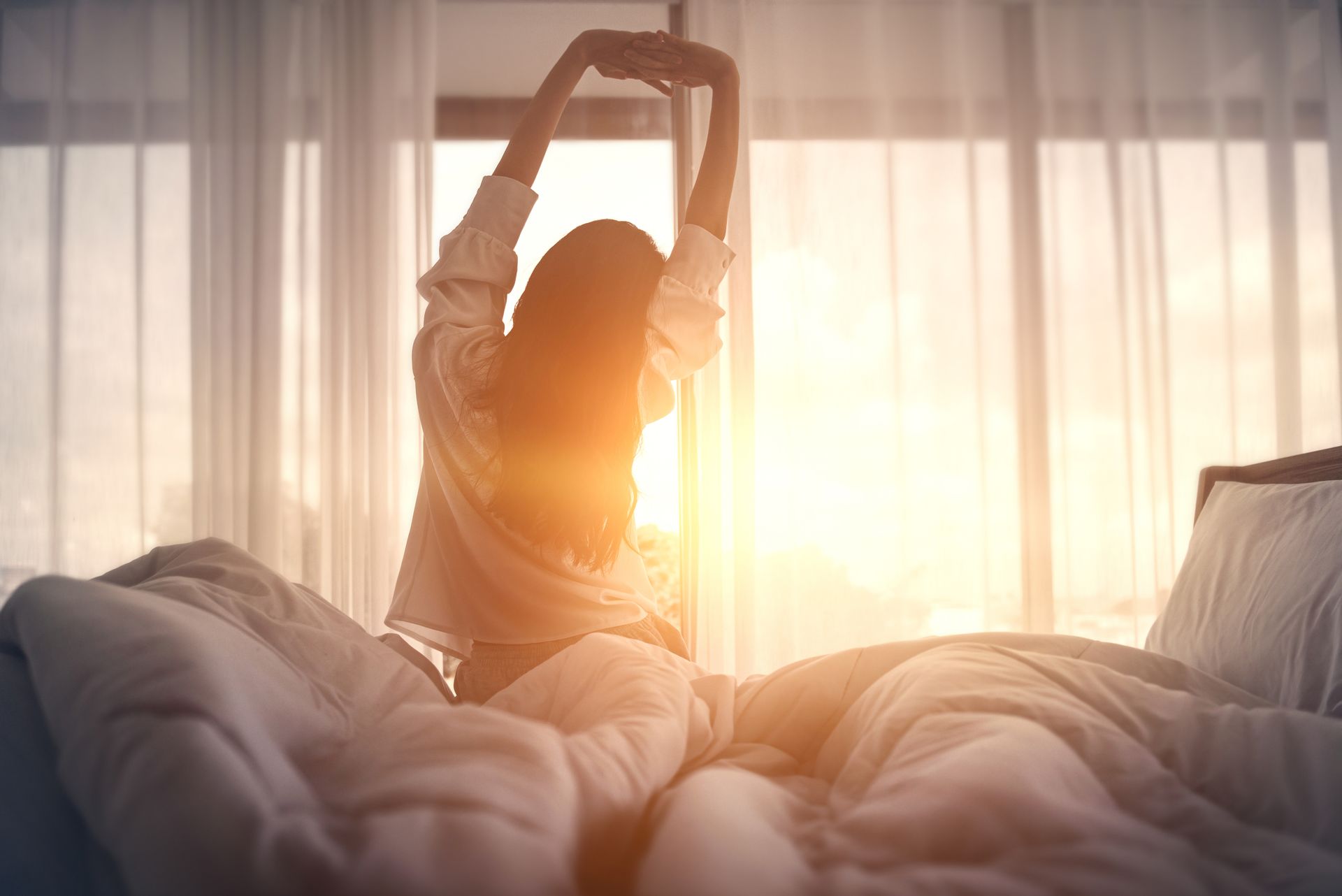 Woman stretching arms above head in bed, with morning sun shining in the east-facing window