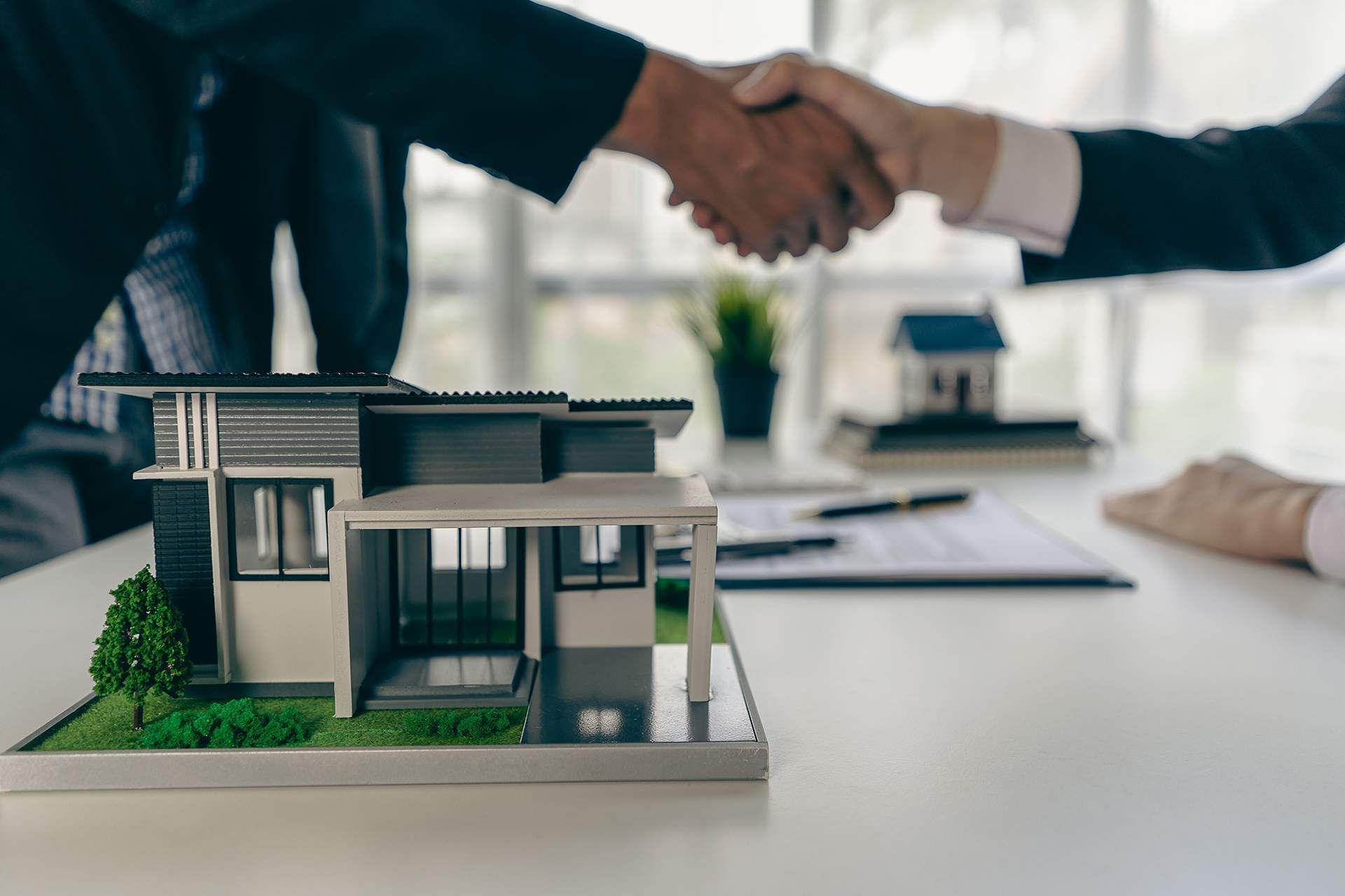 Two people shaking hands over a model house on a desk, with paperwork in the background