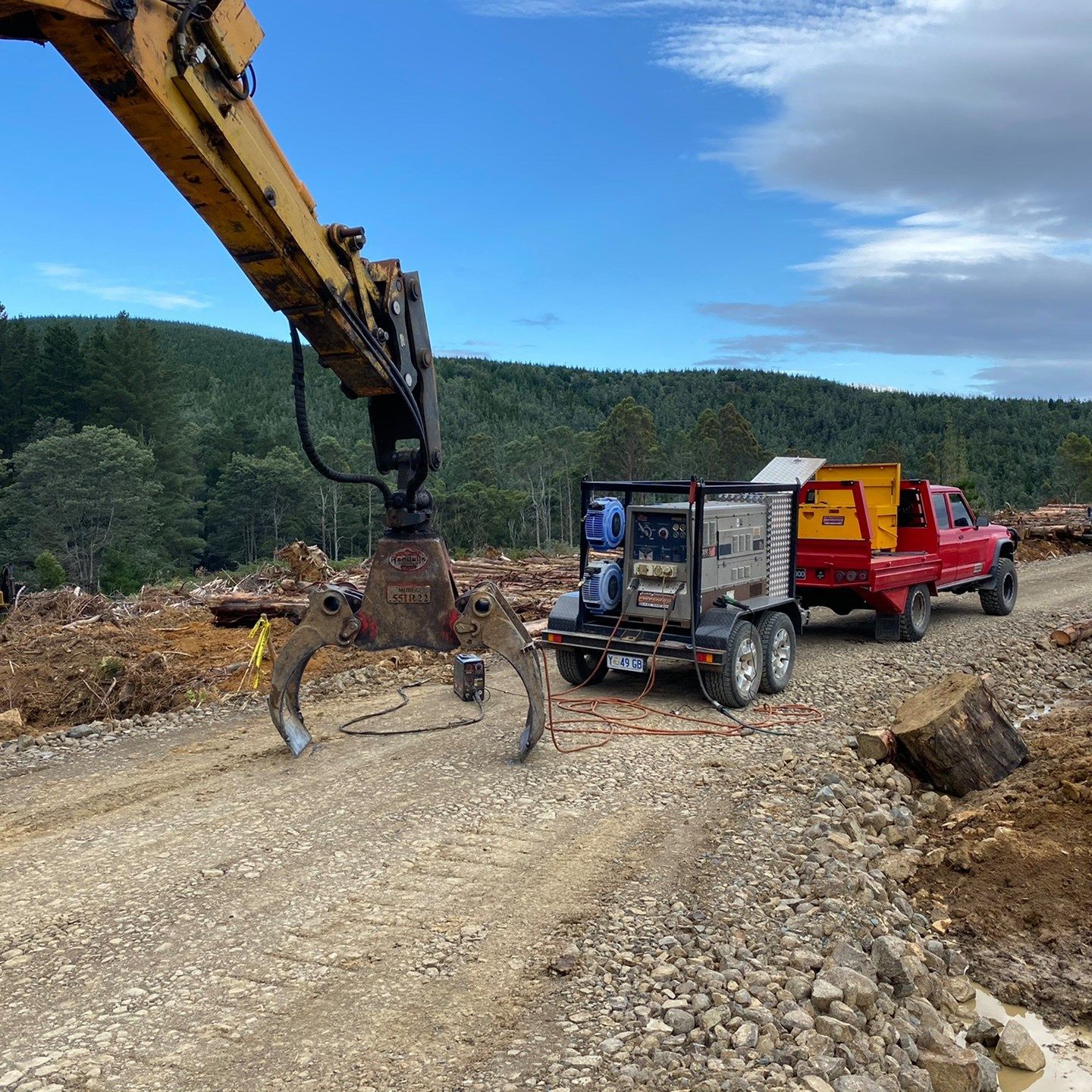 Excavator with grapple near dump truck and red pickup on a gravel road in a cleared forest area
