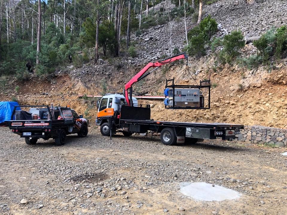 Red crane truck beside a flatbed truck lifting cargo on a gravel road in a wooded hillside area