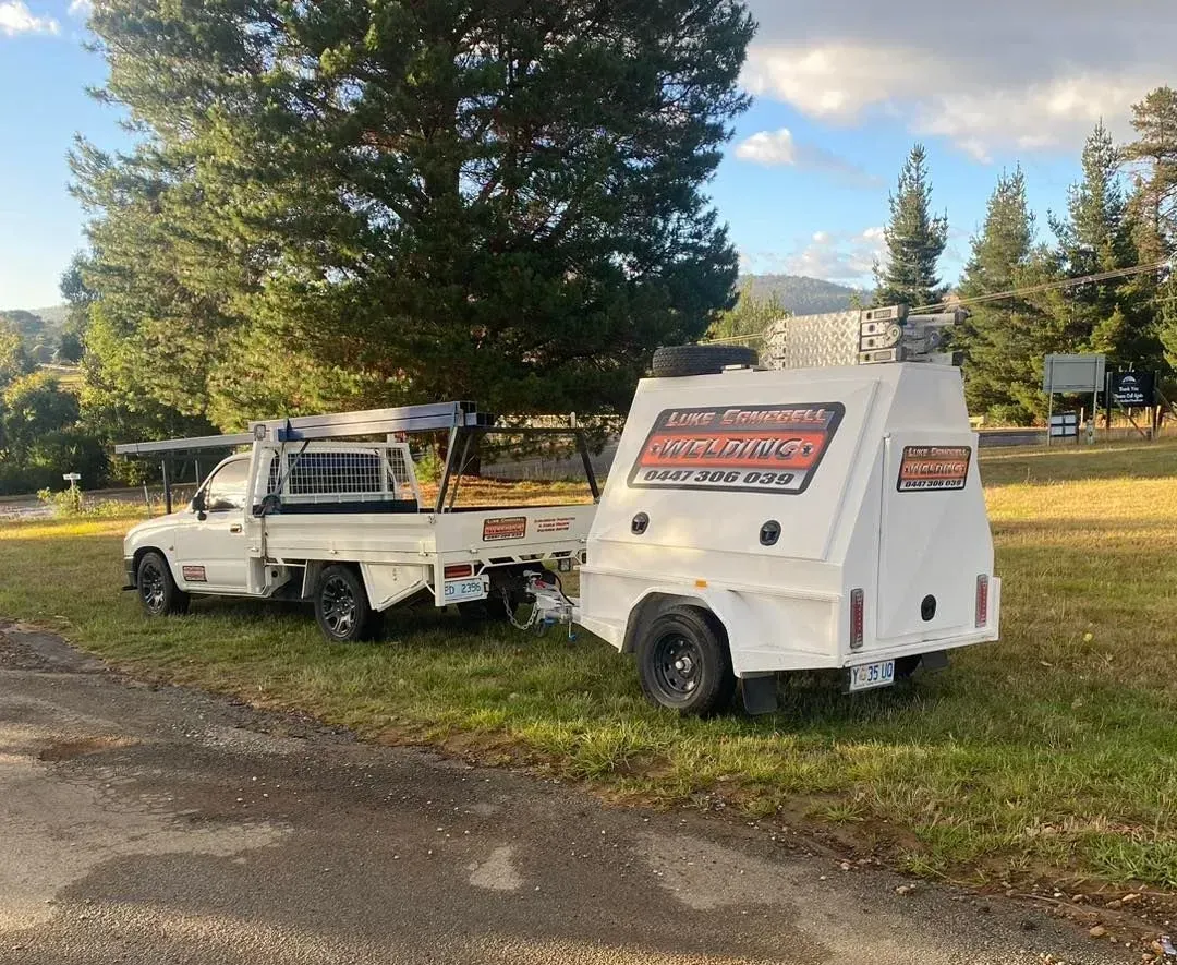 White utility truck with a box trailer parked on grass beside a road, trees in the background.