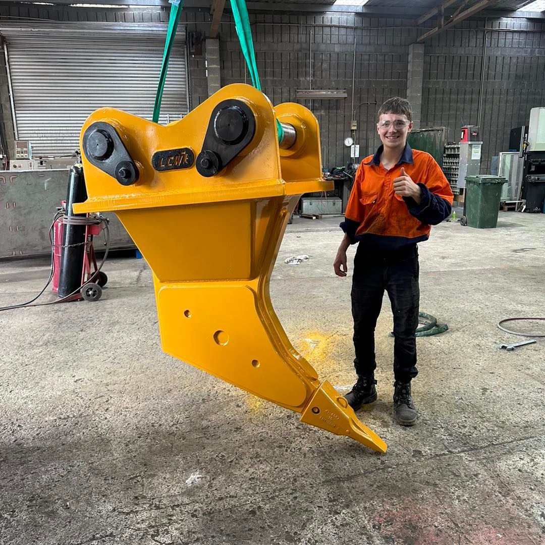 Man in orange shirt standing beside a large yellow excavator attachment in a warehouse