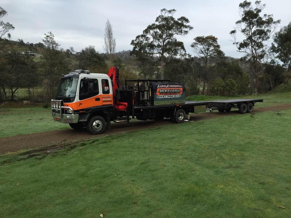 Orange tow truck hauling a flatbed trailer across a grassy field with trees in the background