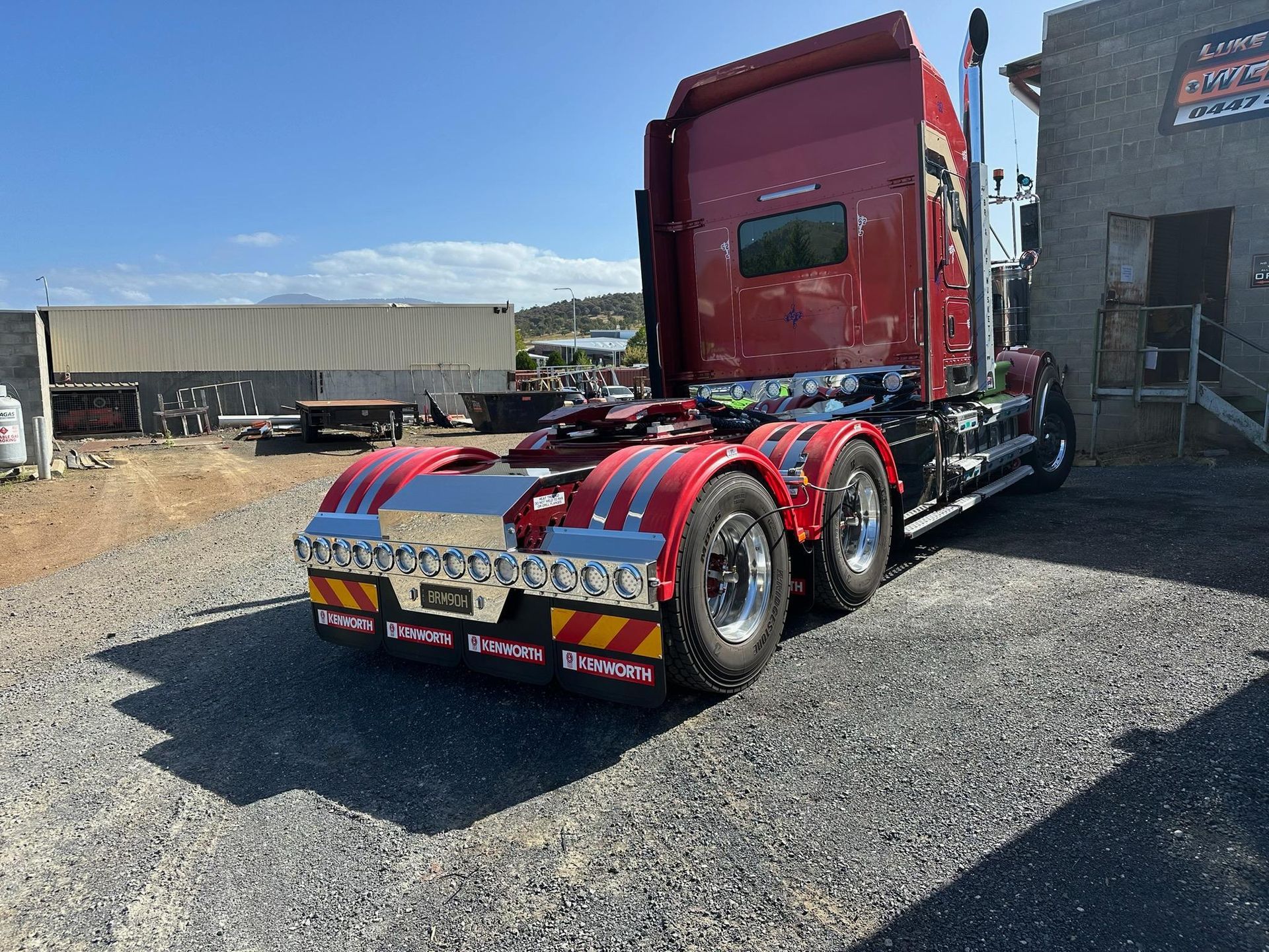 Red semi-truck cab with chrome and red-white stripes parked on gravel in an industrial yard.