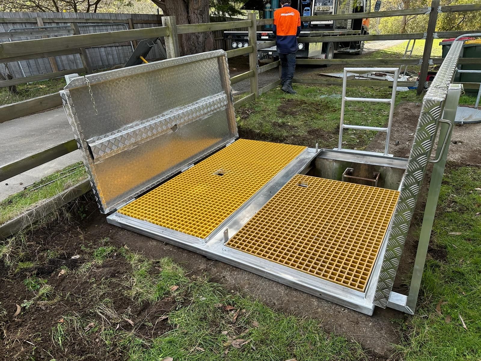 Metal livestock ramp with yellow grated platforms in a fenced outdoor area, worker in orange jacket nearby