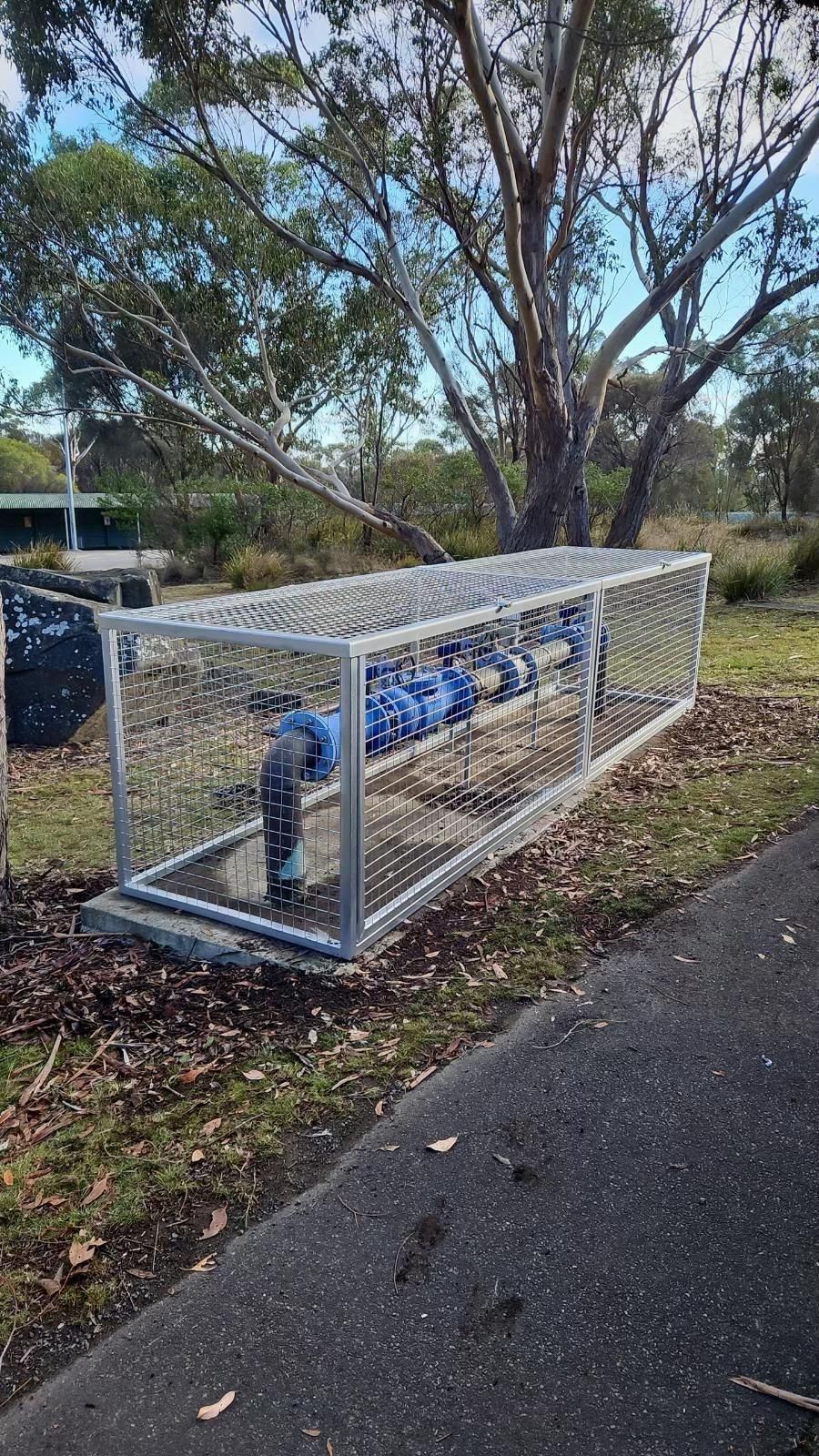 Blue metal pipe beside a paved path, enclosed in white chain-link fencing under trees.