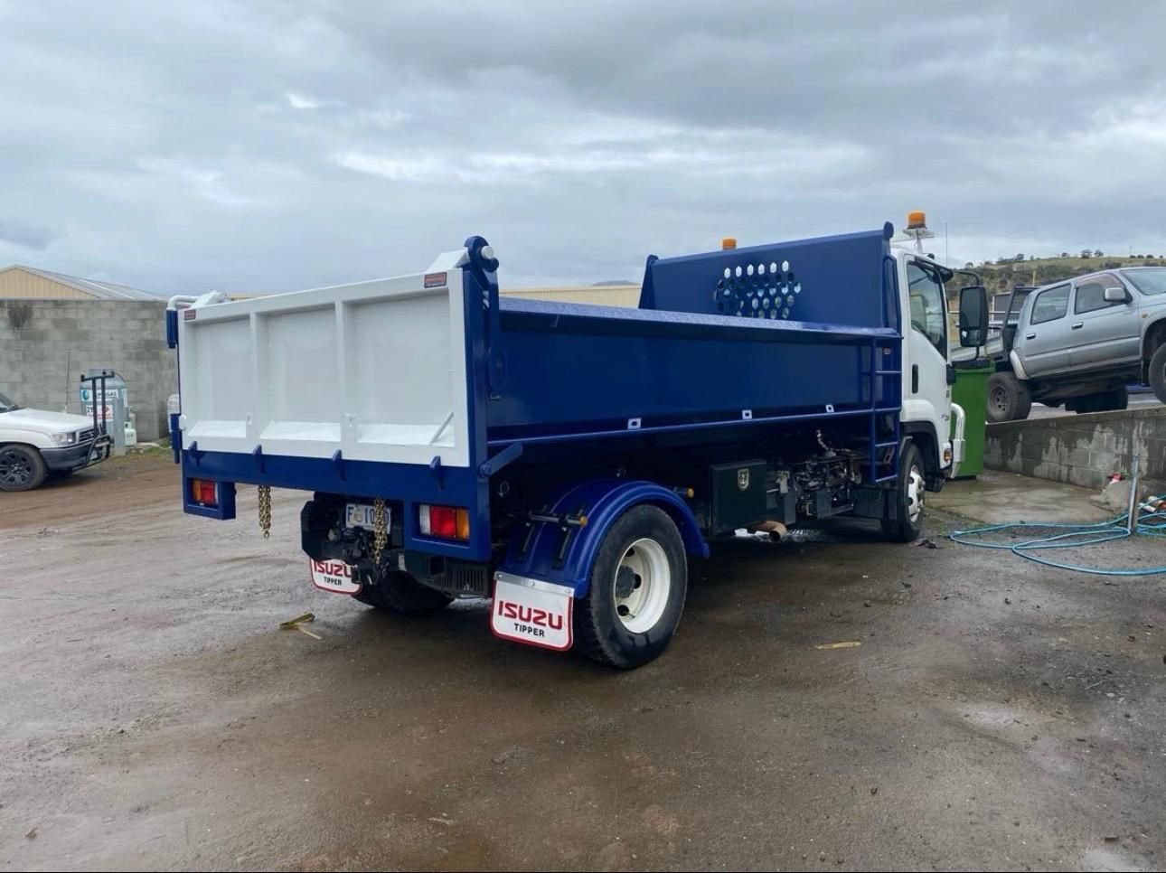 Blue dump truck on a muddy lot under a cloudy sky, viewed from the rear side.