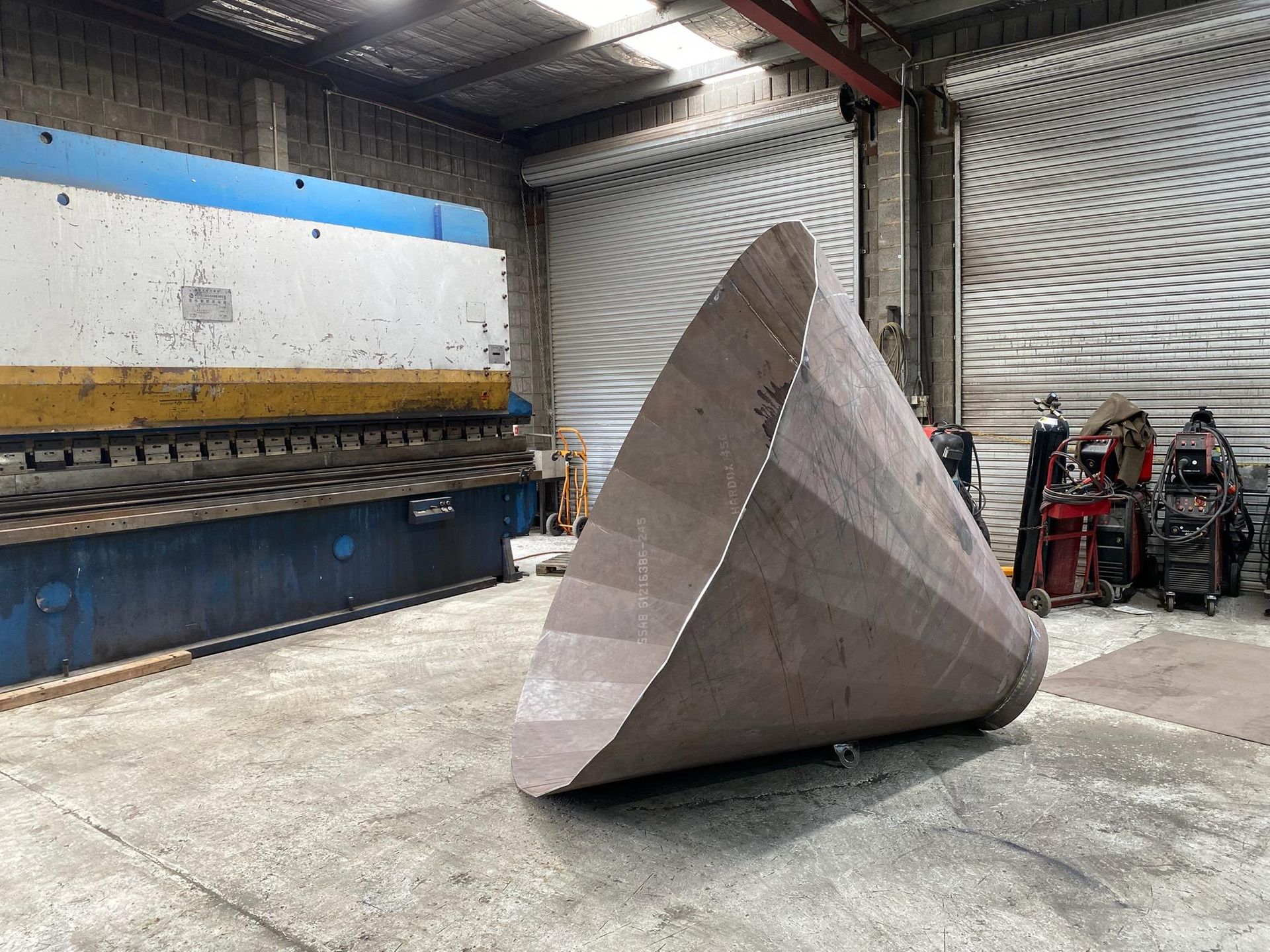 Large triangular metal sculpture in an industrial warehouse, with a blue and yellow machine behind it.