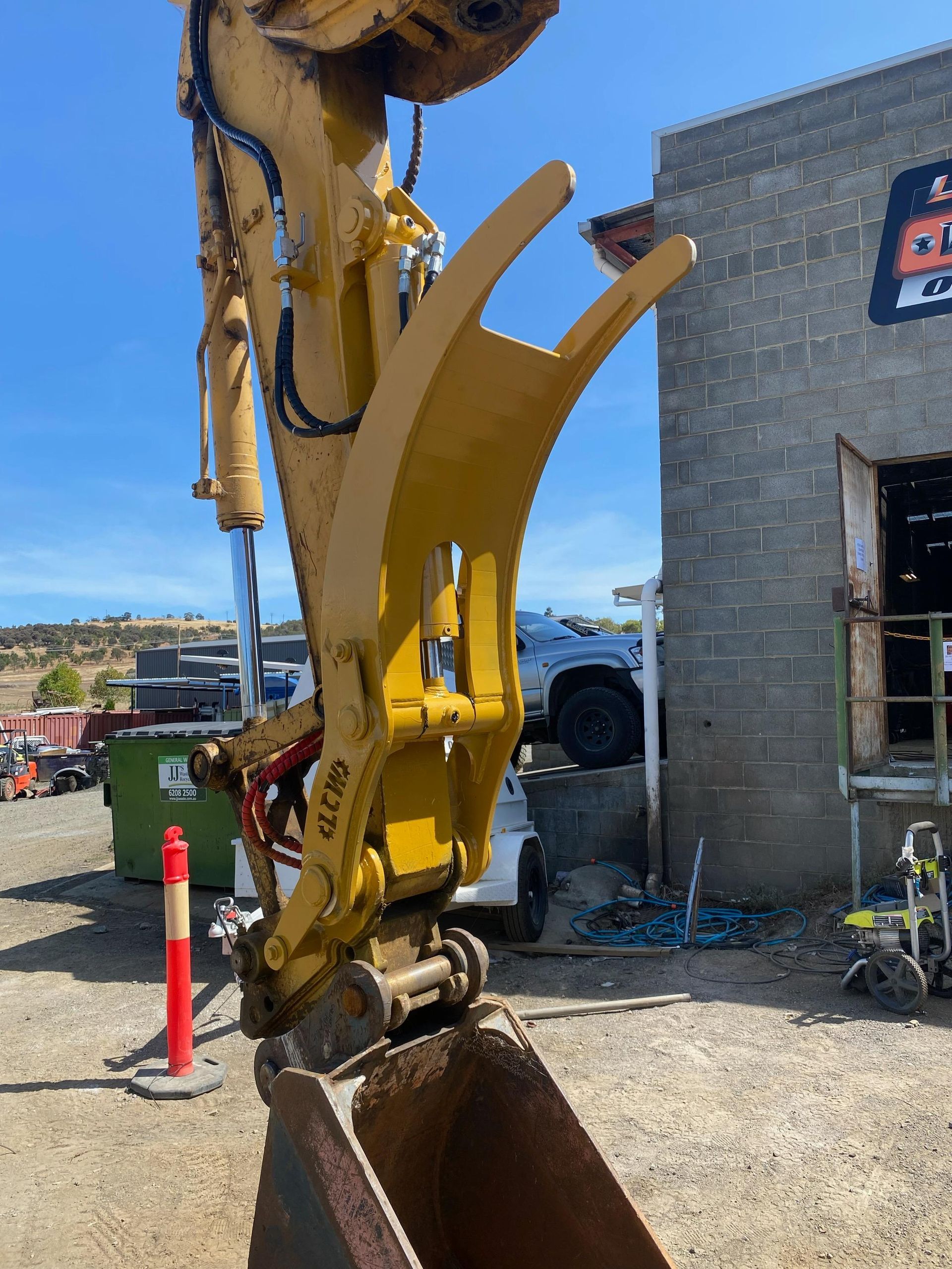 Yellow excavator bucket beside a brick building at a construction site, with gravel and orange cones.
