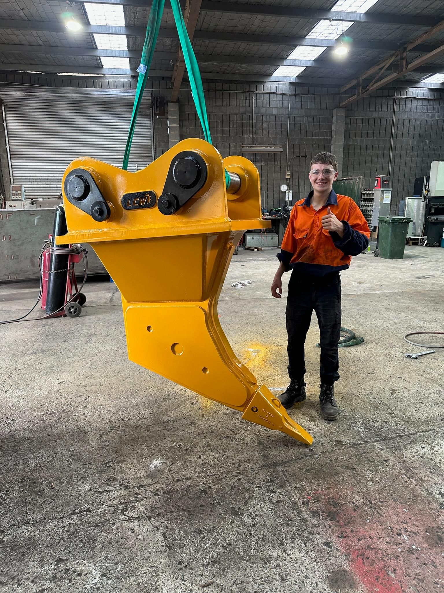 Yellow excavator bucket suspended by green straps in a warehouse, with a man in orange standing beside it