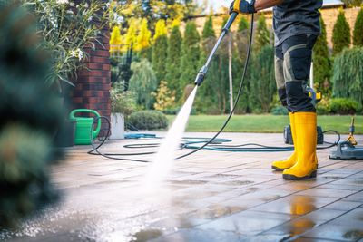 Person pressure-washing a driveway with yellow boots and a green hose on a sunny day