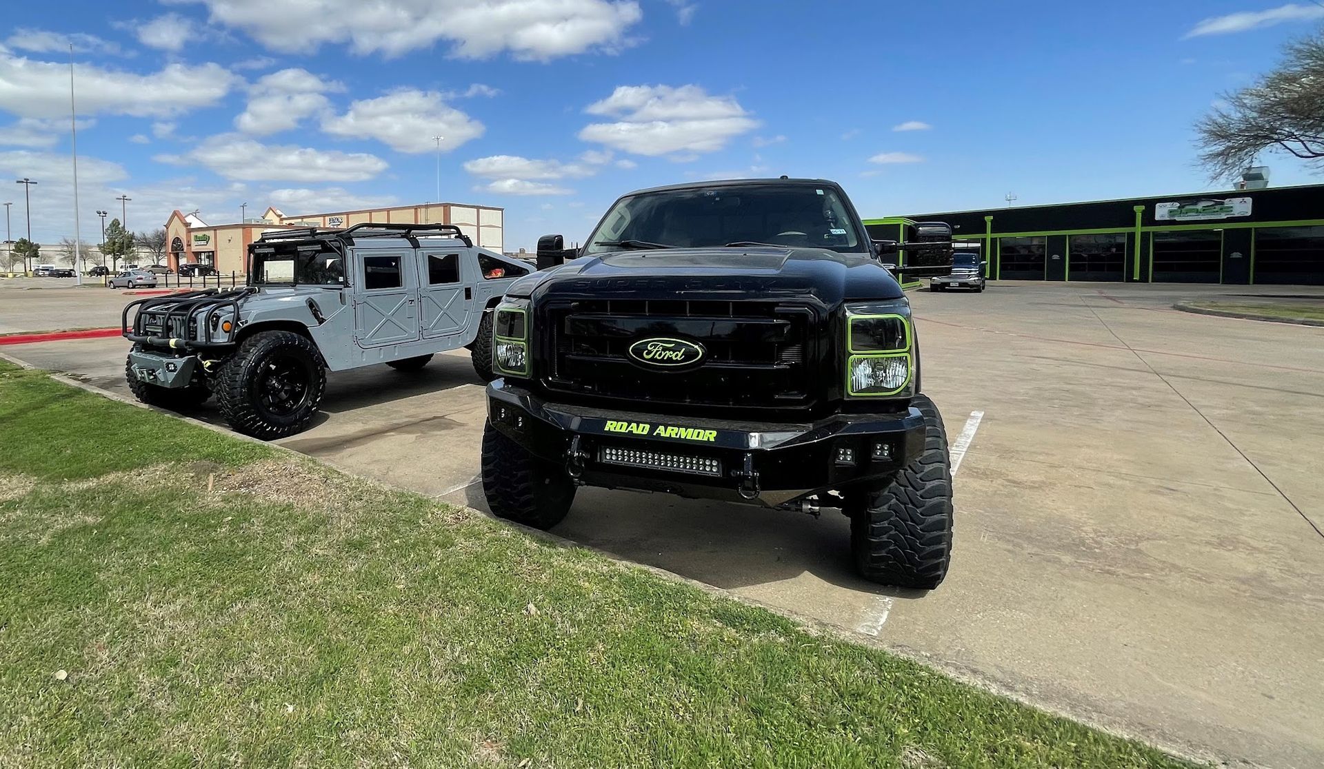 A black lifted Ford pickup truck and a gray Hummer parked on a paved area with a grassy area in front, under a cloudy sky.