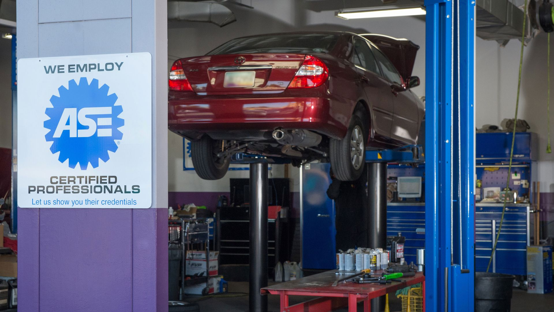 Red car lifted on a hydraulic auto shop lift inside a garage with tools and equipment nearby