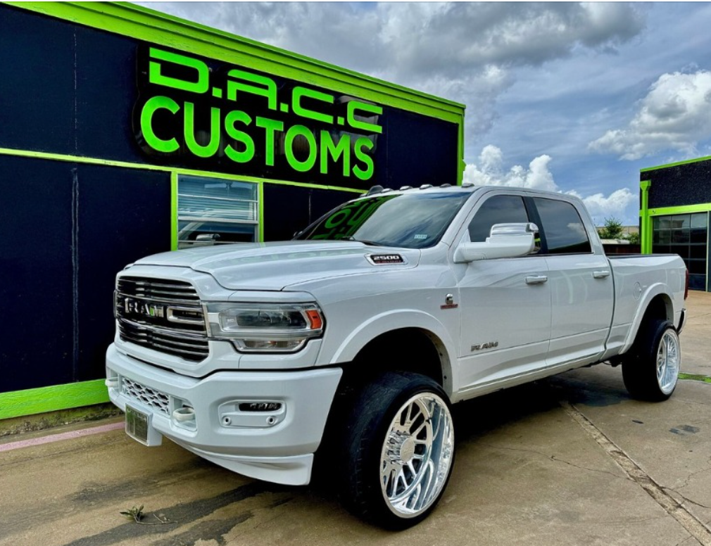 White Ram truck parked in front of D.R.C.C. Customs shop with a lime green logo. The truck has large chrome wheels.