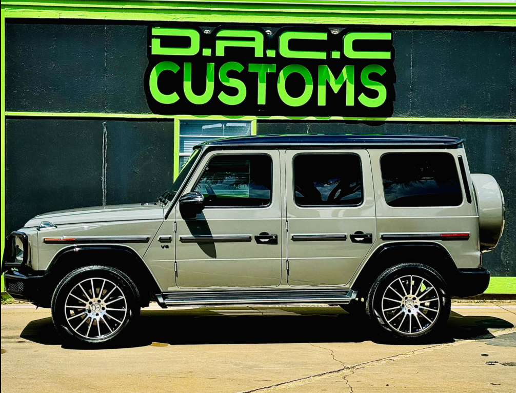 A silver Mercedes G-Wagon parked in front of a custom shop with a green and black sign, 
