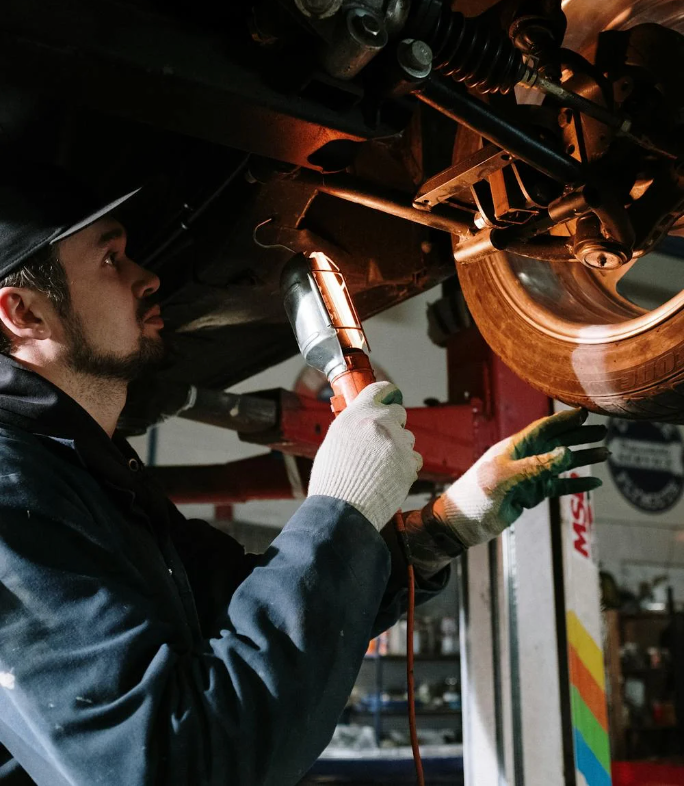 Mechanic wearing gloves examines car undercarriage with a spotlight. He’s in a garage, looking at a tire.