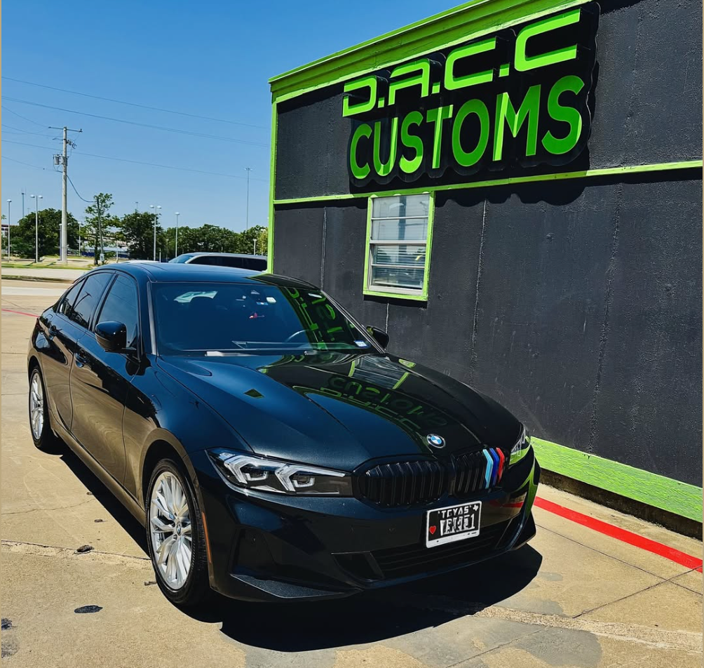 Black BMW sedan parked in front of D.A.C.C. Customs with green and black signage on a sunny day.