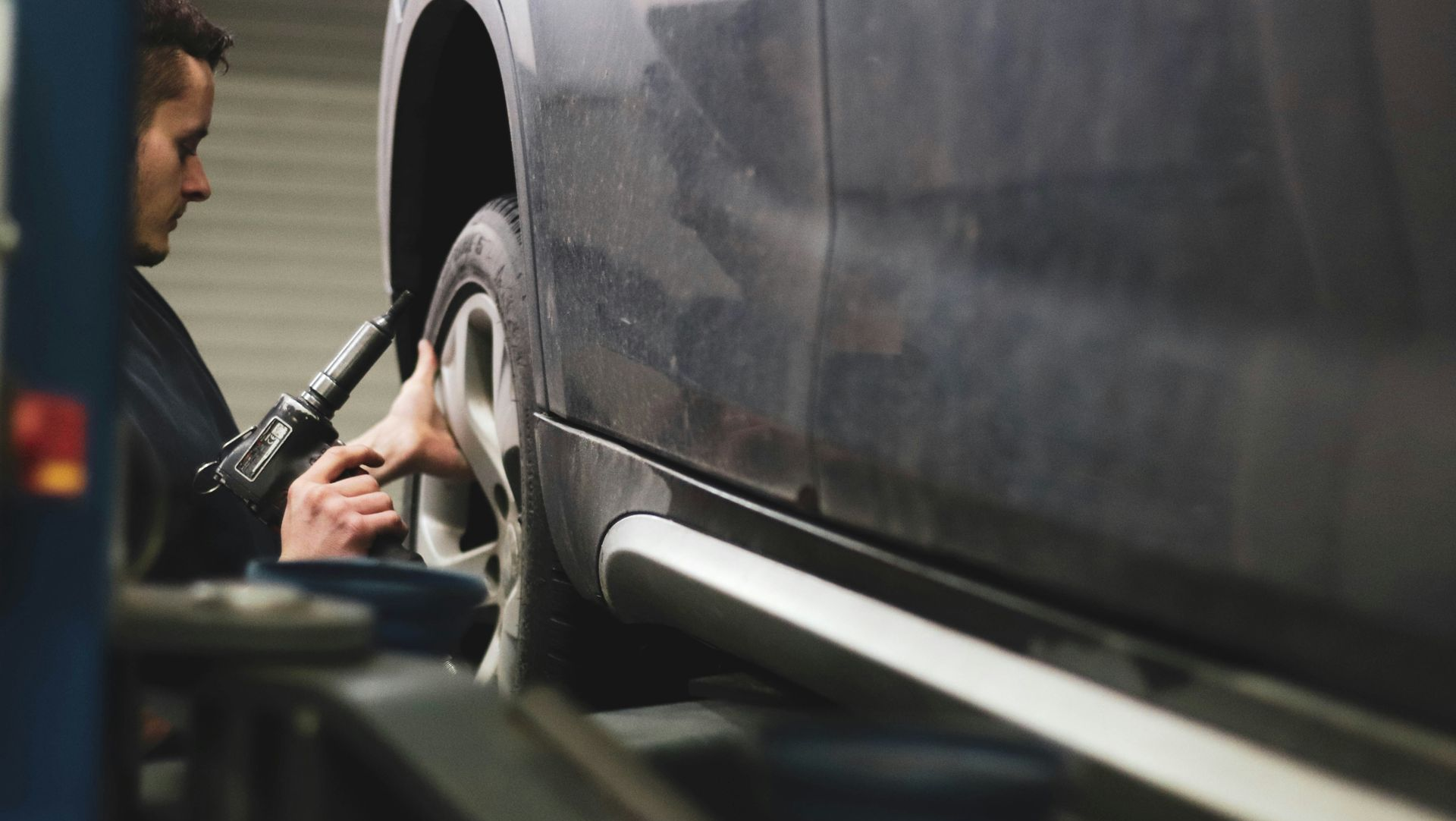 Mechanic inspecting a car tire with a tool beside a dark vehicle in a repair bay
