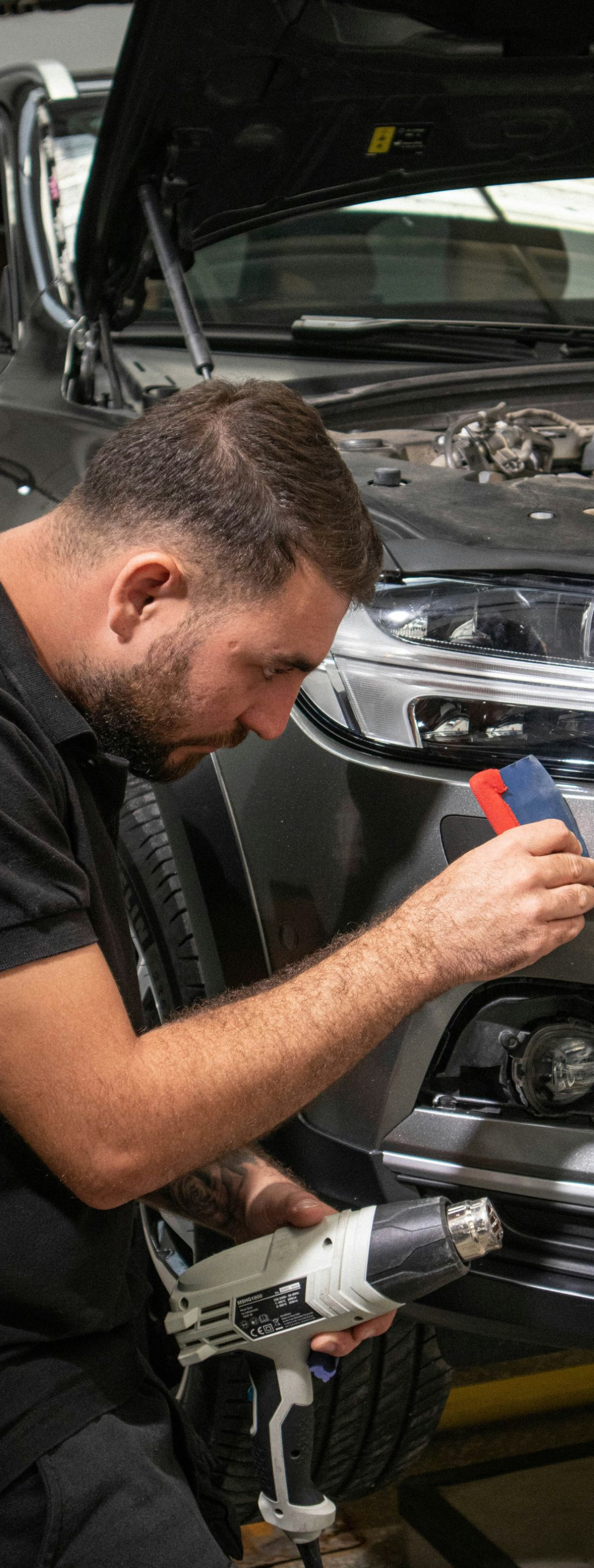 Mechanic using a power drill to repair the front bumper of a gray car in a garage