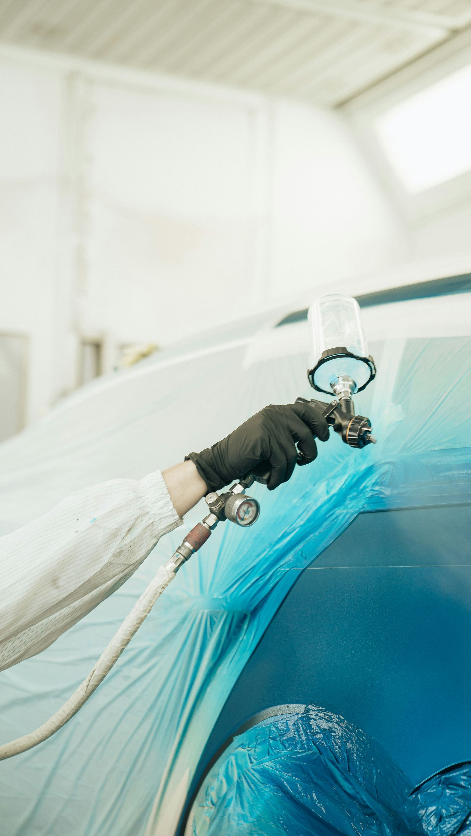 A gloved hand uses a spray paint gun to apply paint to a blue-masked vehicle component in a workshop.