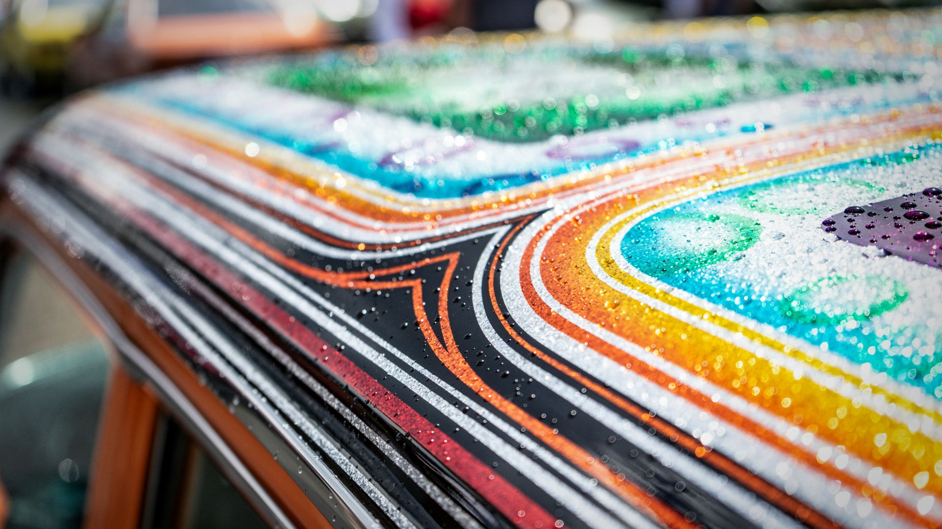 Close-up of a car roof with intricate, colorful, glittery paint patterns and pinstriping, covered in tiny water droplets.