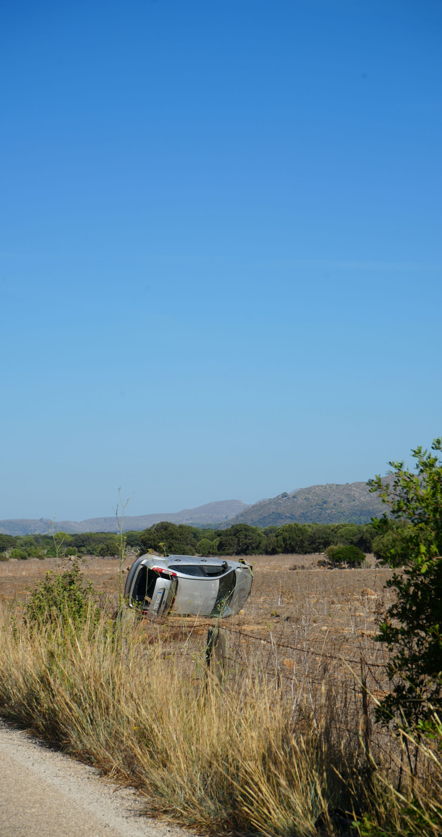 A silver car flipped upside down in a dry, rural field next to a dirt road under a clear blue sky.