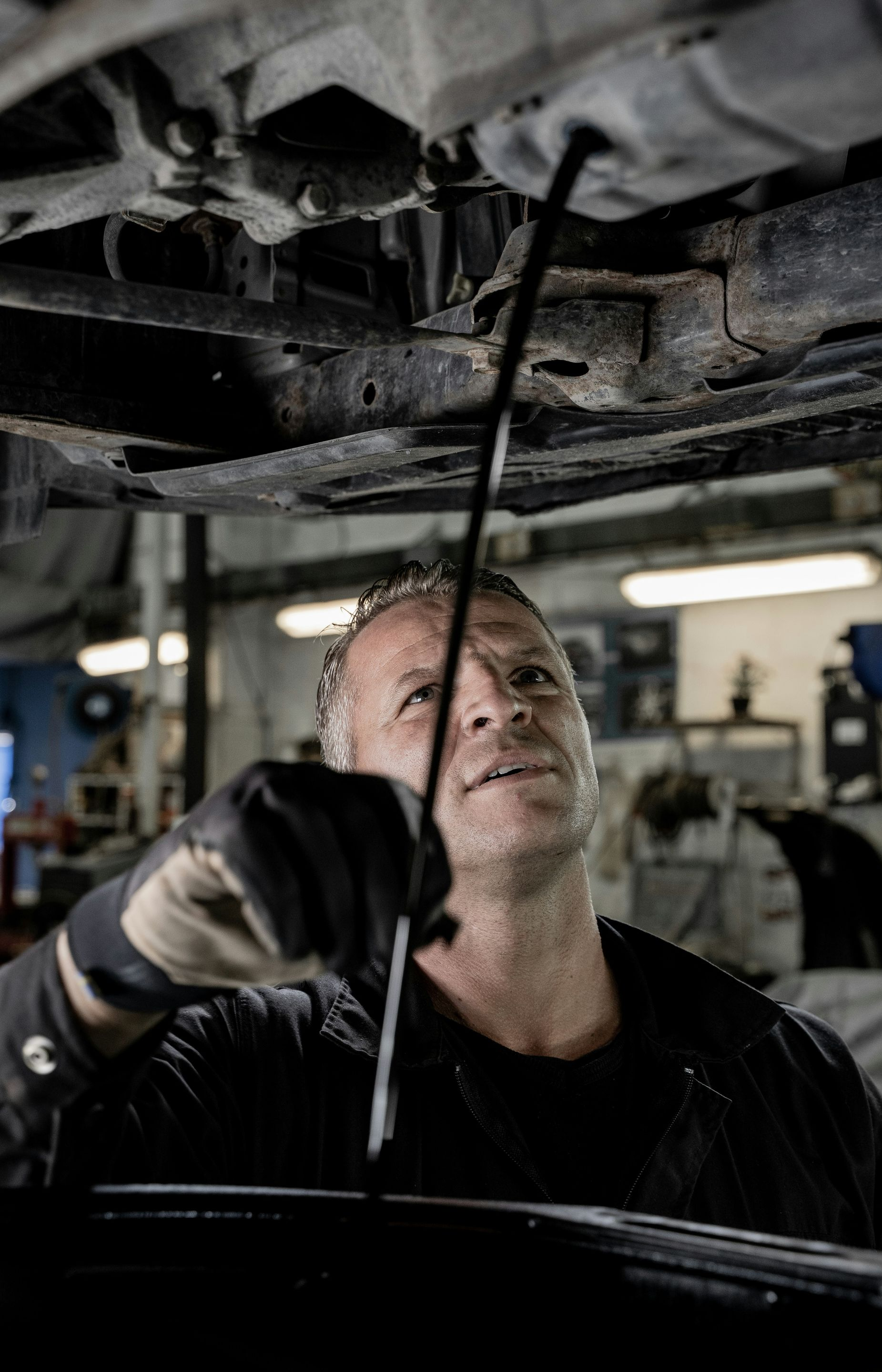 A mechanic wearing gloves examines a stream of dark oil draining from the undercarriage of a vehicle in a repair shop.