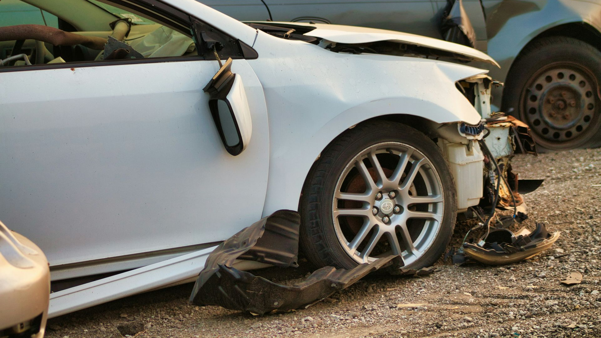 Damaged white car with crushed front end after a collision, parked beside another vehicle on gravel.