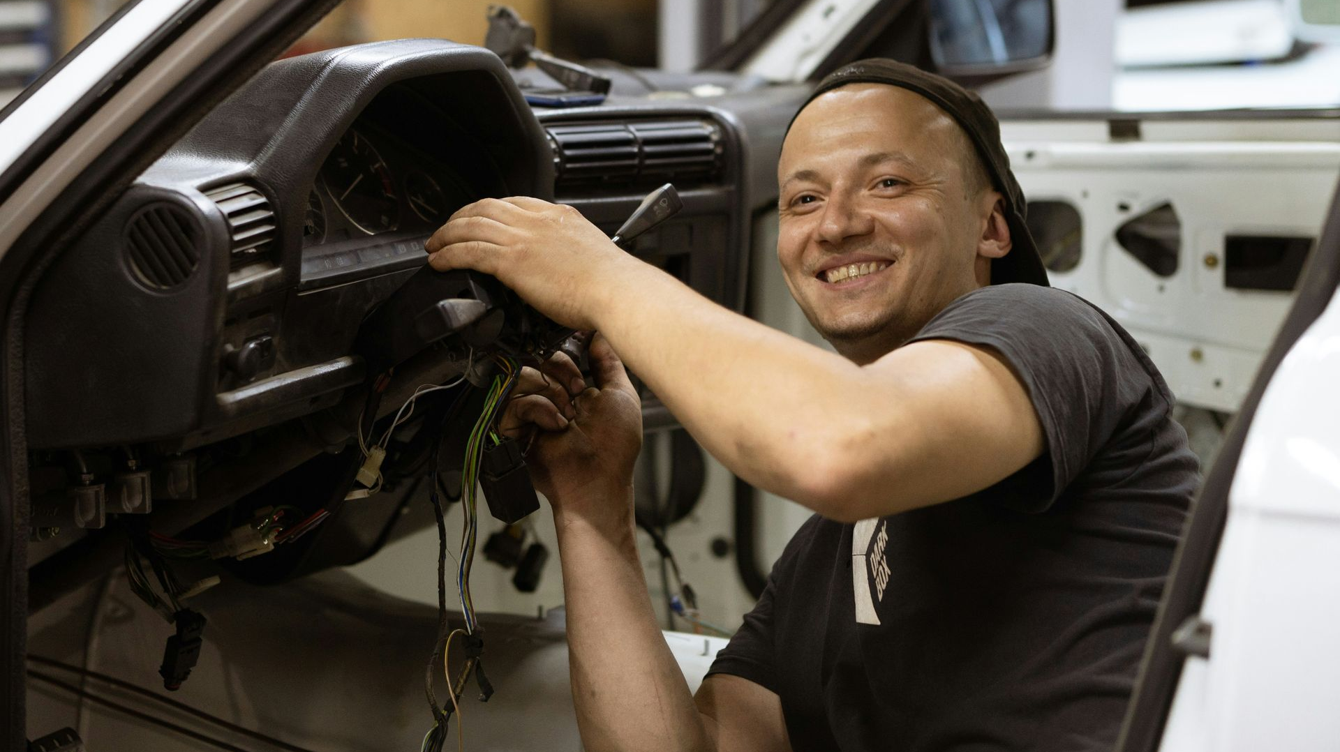 A person wearing a beanie smiles while working on the exposed steering column and dashboard of a car.