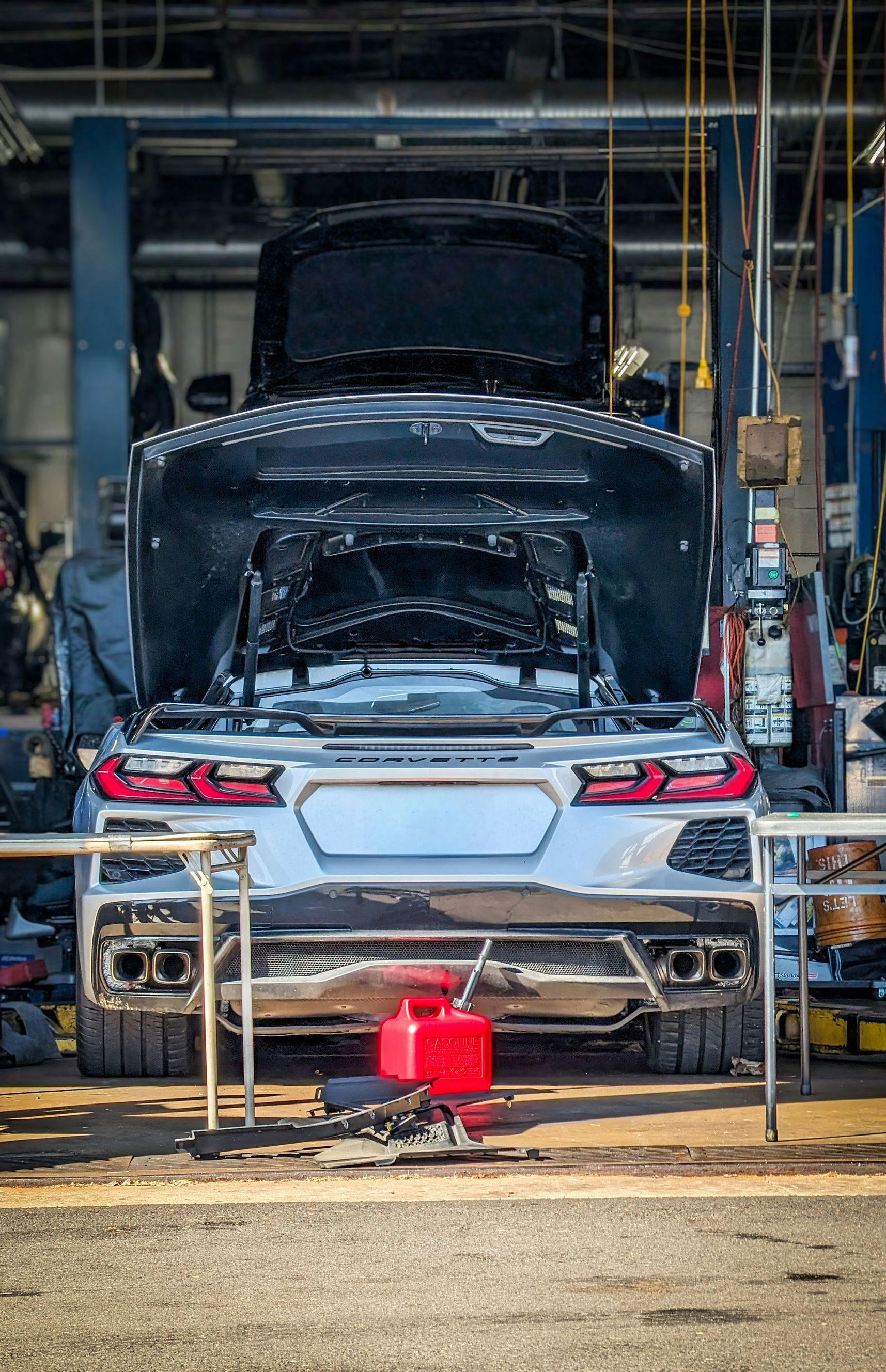 A black sports car in a repair shop with its hood open and rear bumper removed, surrounded by tools.