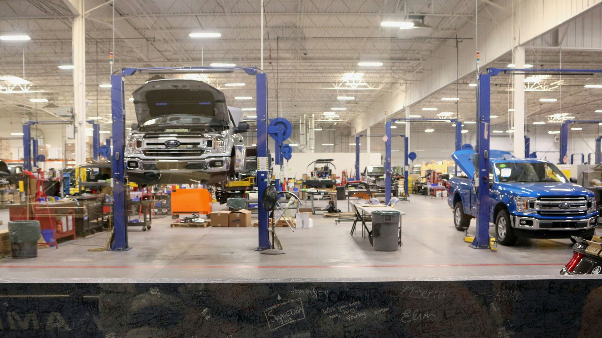 A brightly lit auto repair shop featuring a lifted silver pickup truck and a blue pickup truck parked on the floor.