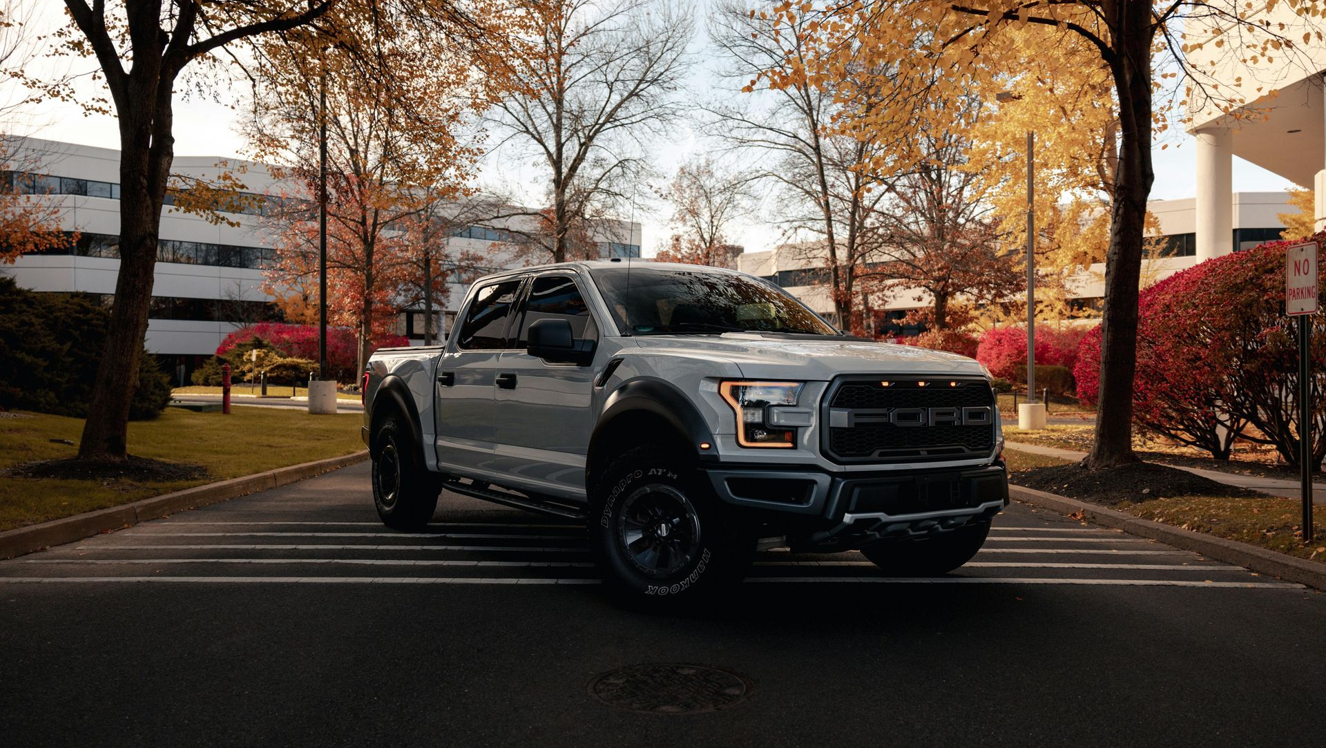 Gray pickup truck parked on a paved road lined with autumn trees near office buildings