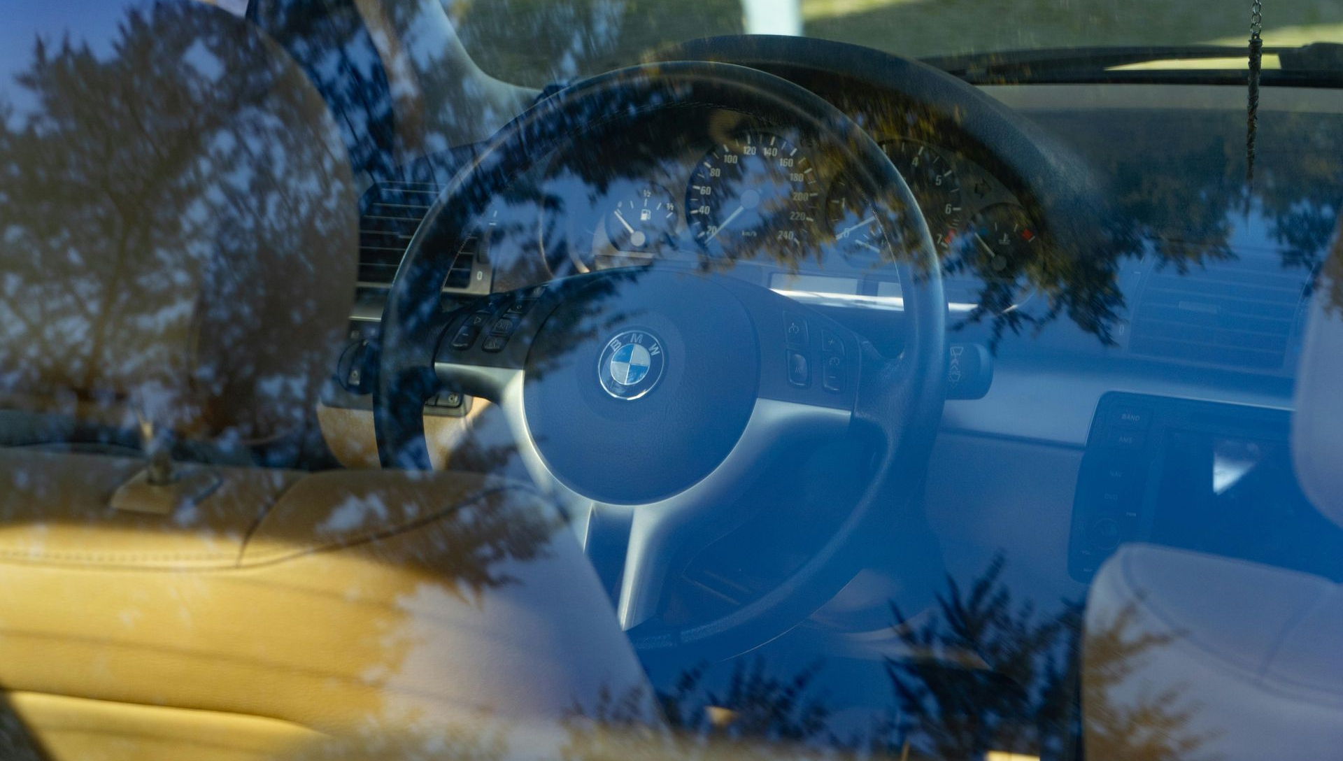 Steering wheel of a BMW car, viewed through a windshield with tree reflections.