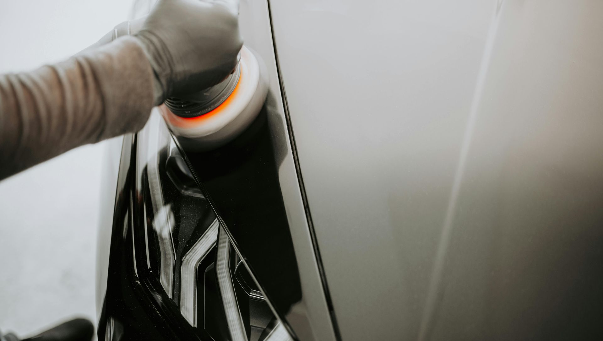 A person wearing a glove uses a power buffer tool to polish the headlight of a white car.