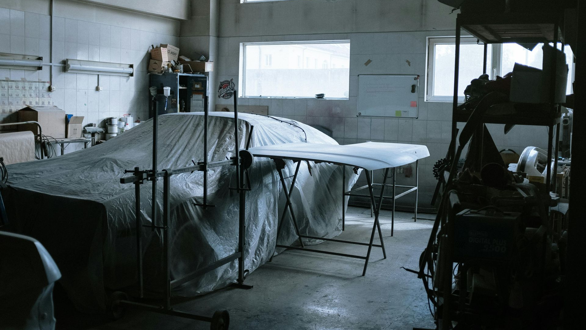 A car covered in plastic sheeting inside a dim, industrial workshop with shelving units and light streaming from windows.