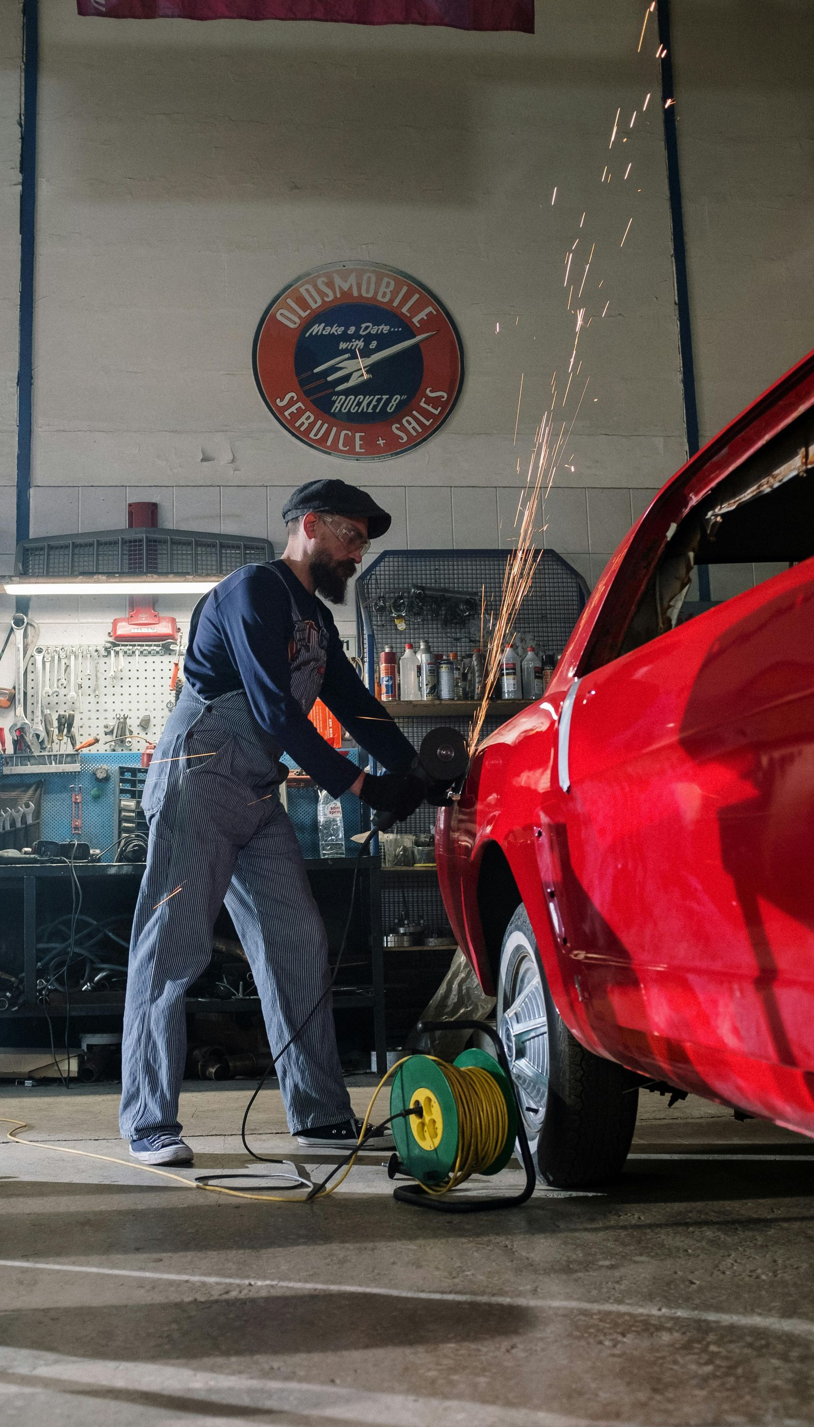 Mechanic in striped overalls using a grinder on a red car in a garage, sparks flying.