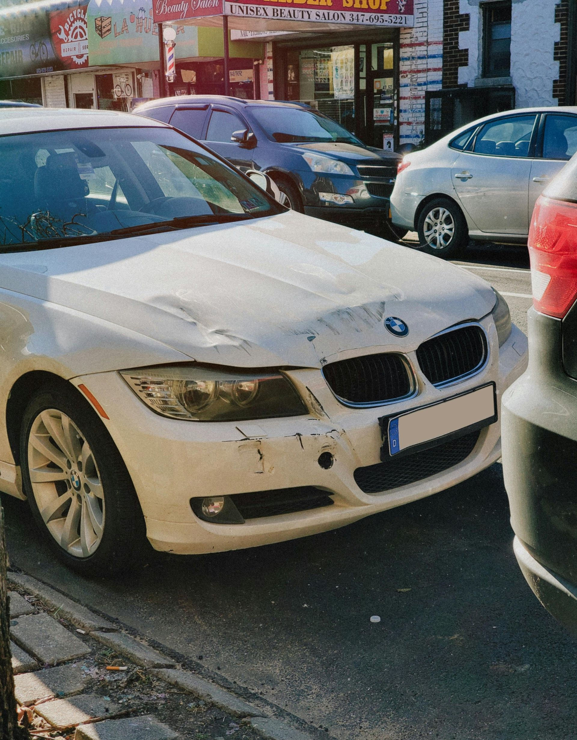 White BMW car with front-end damage parked on a city street; other cars and a building in the background.