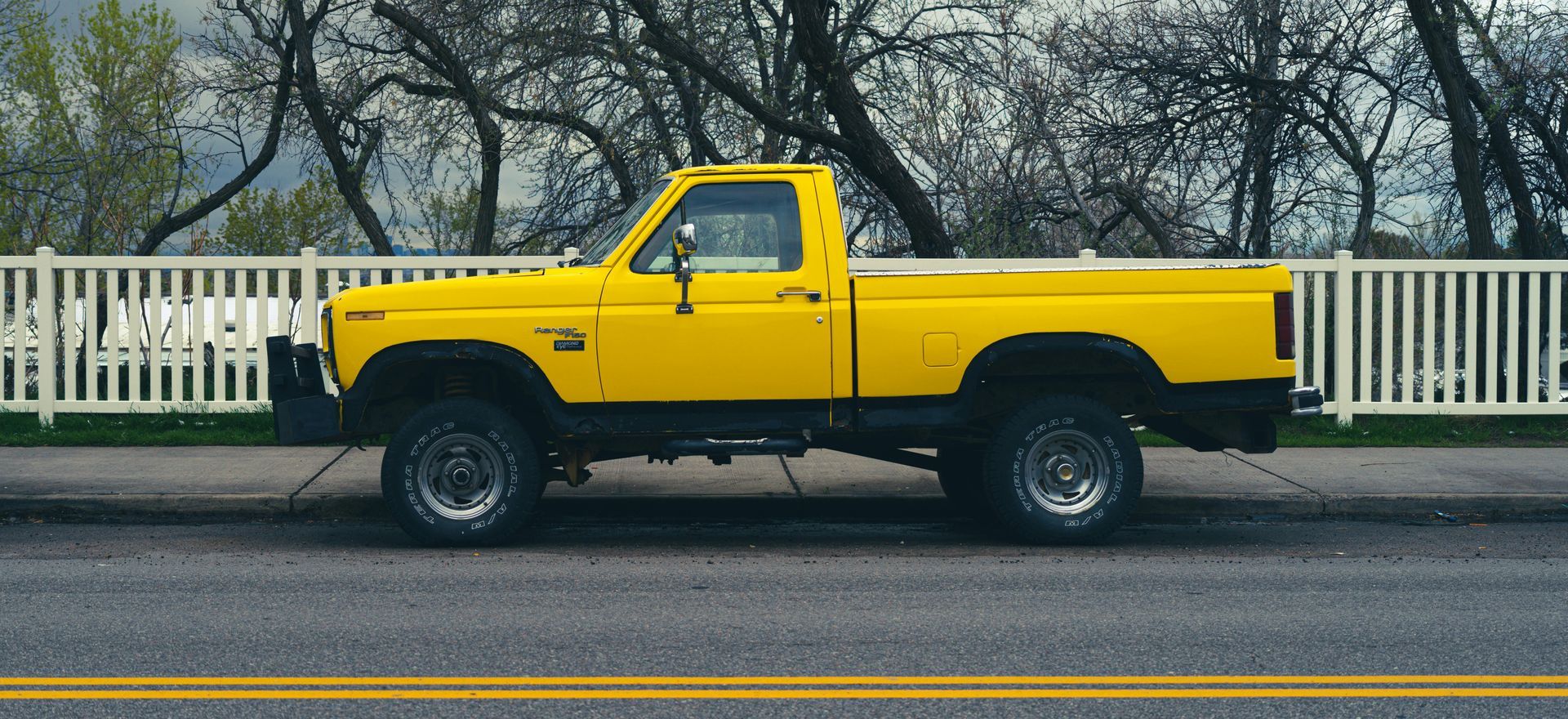 Yellow pickup truck parked on a road beside a white fence with bare trees in the background