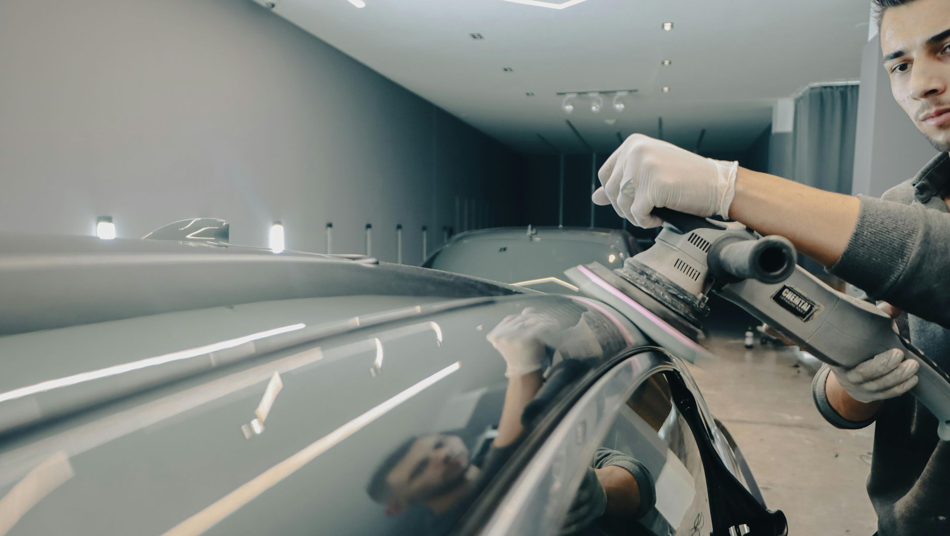 A technician uses a rotary polisher to buff the roof of a car in a brightly lit professional garage.