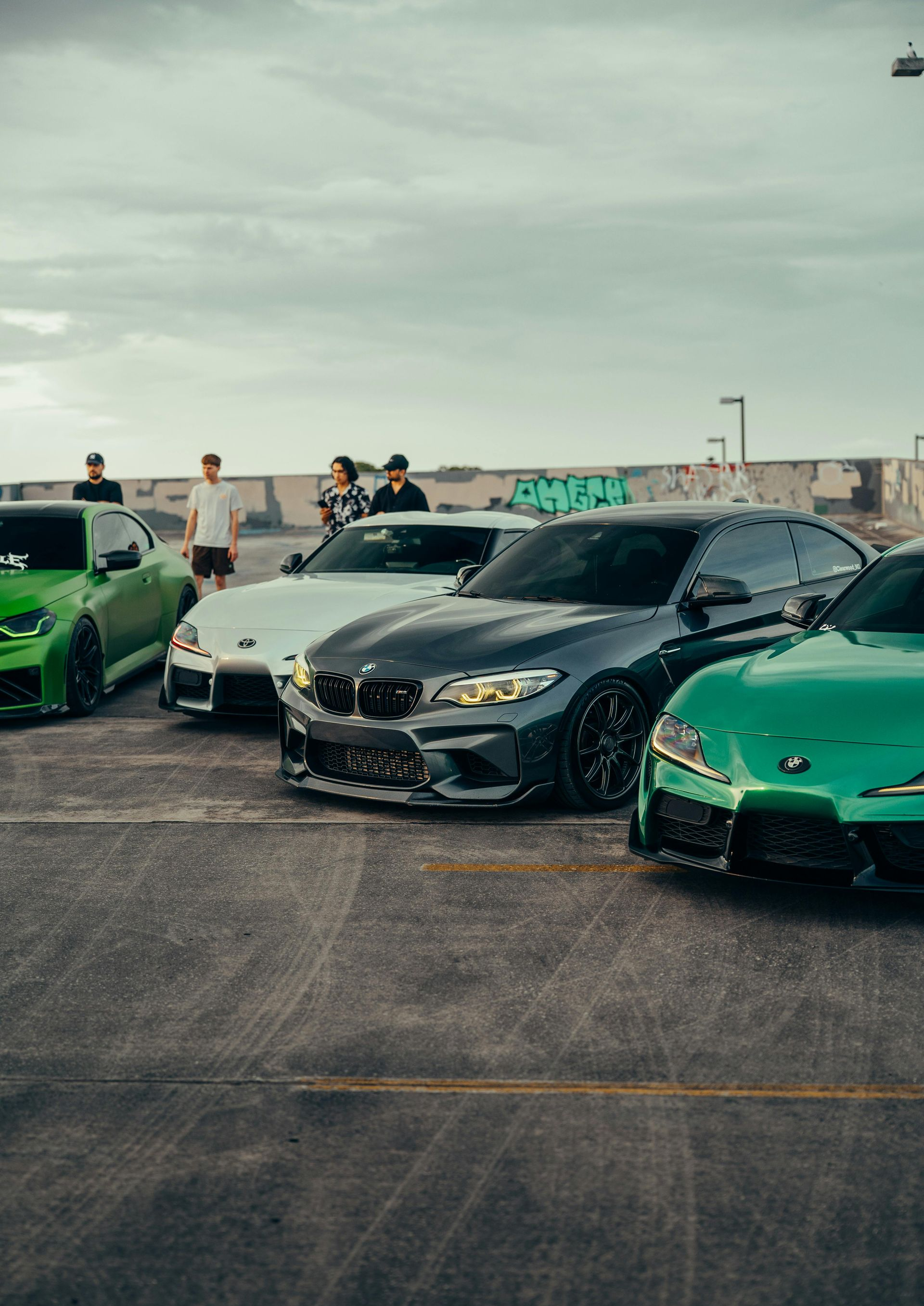 Black BMW sedan flanked by green sports cars at an outdoor car meet under cloudy skies