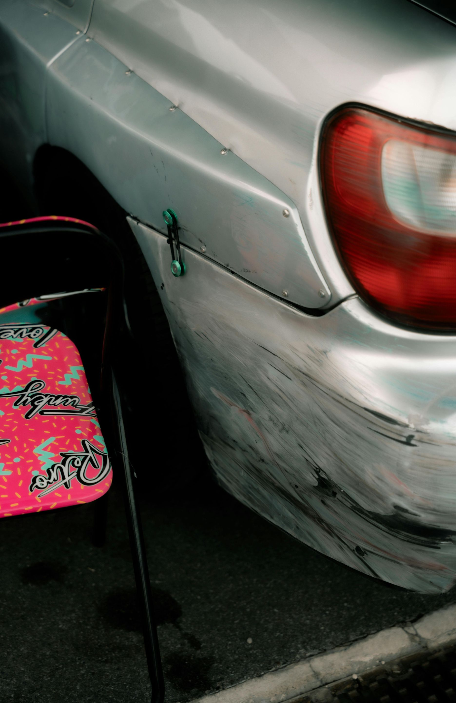 Silver car with damage to the rear bumper, next to a black and pink patterned chair.