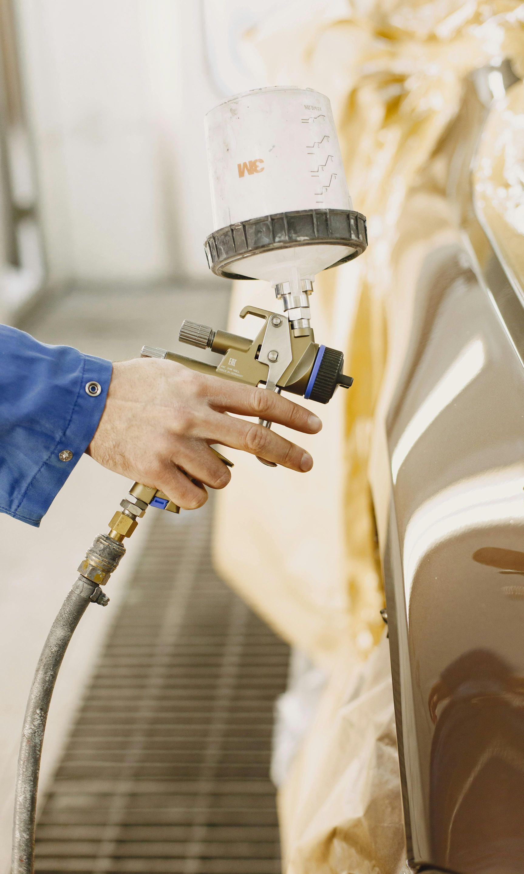 A person in a blue uniform uses a paint spray gun to apply a finish to the side of a car in a workshop.