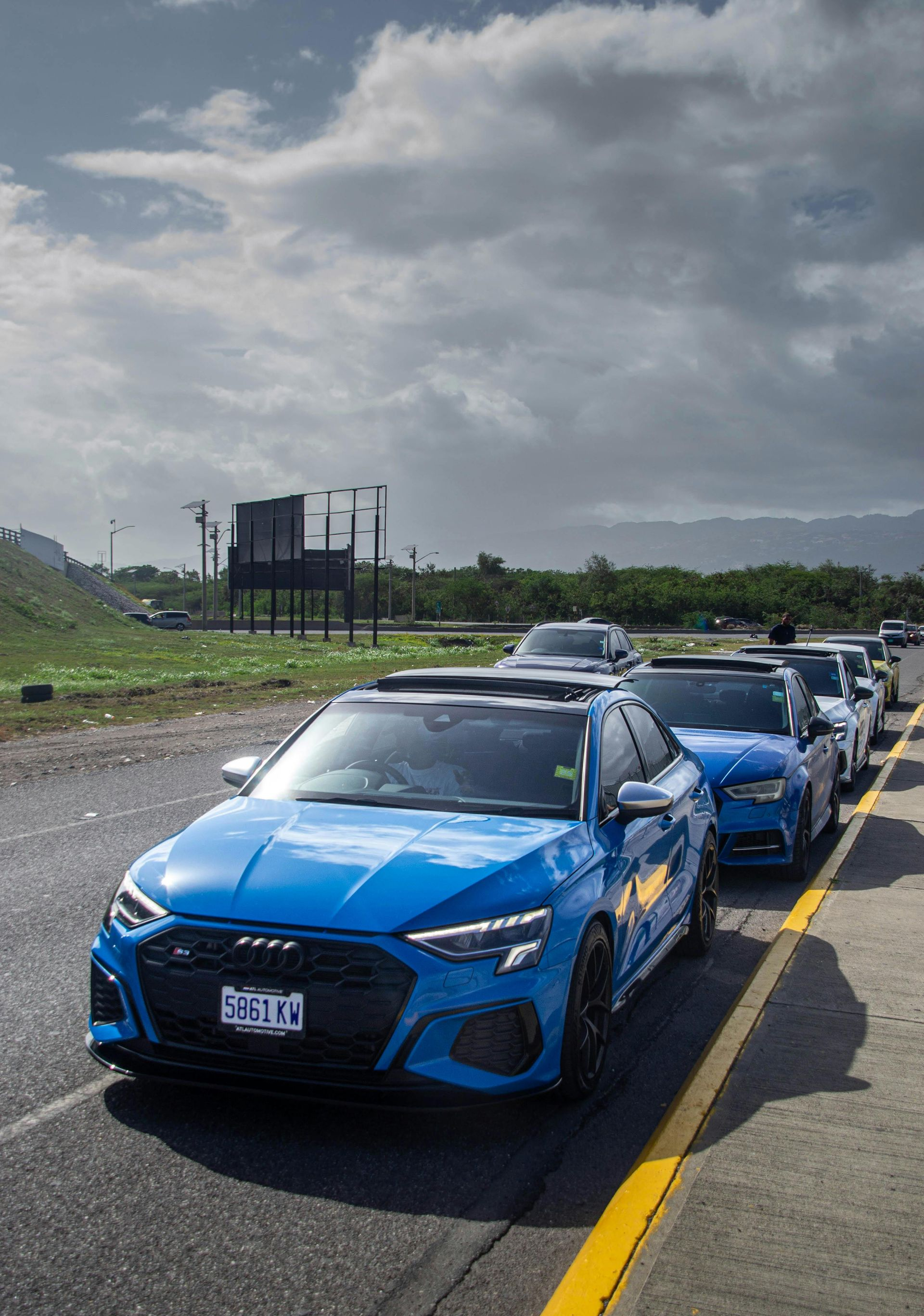 A line of bright blue Audi sedans parked on a roadside under a cloudy sky.