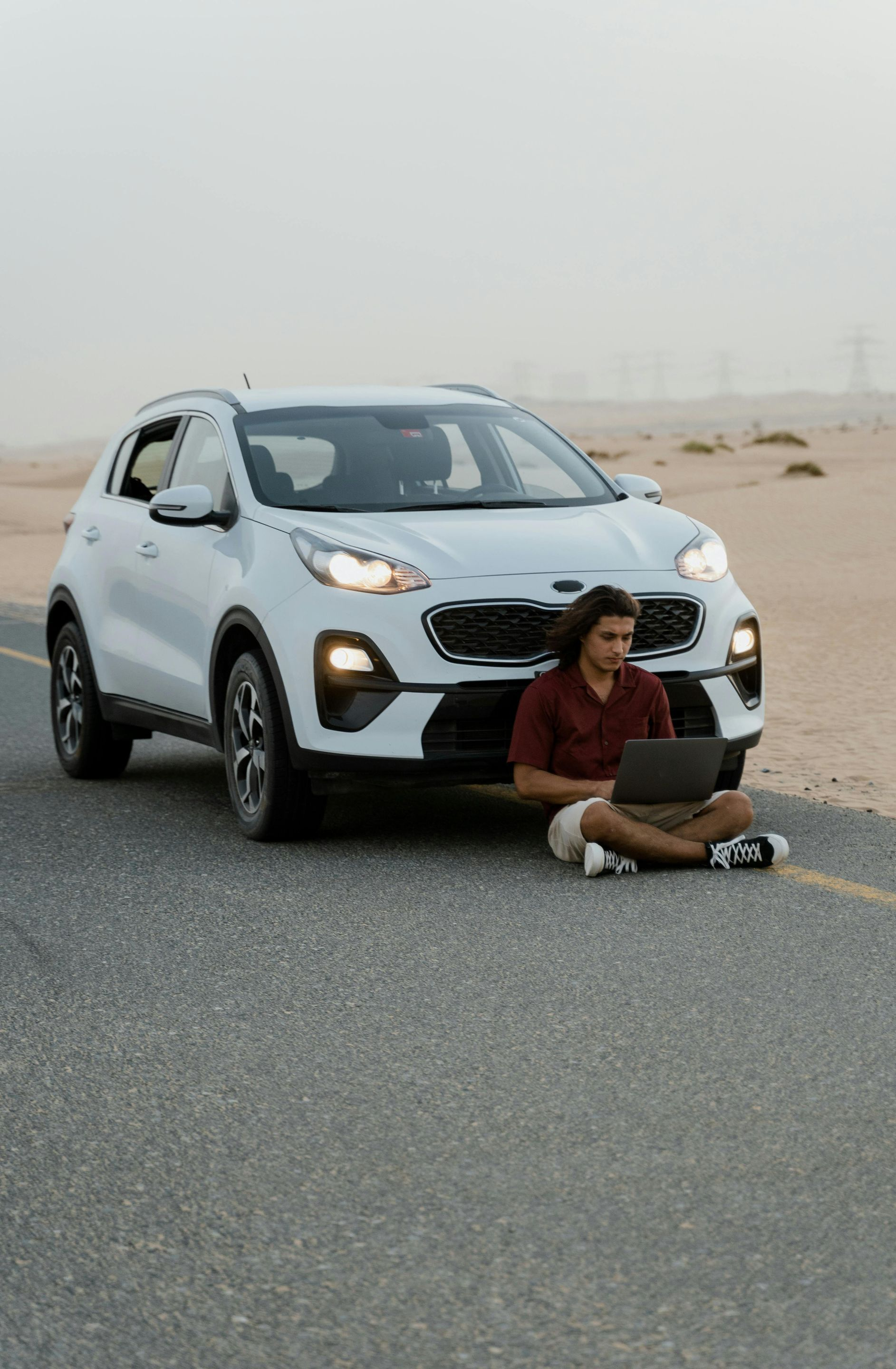 A person sits on an asphalt road in a desert landscape, using a laptop in front of a white SUV.