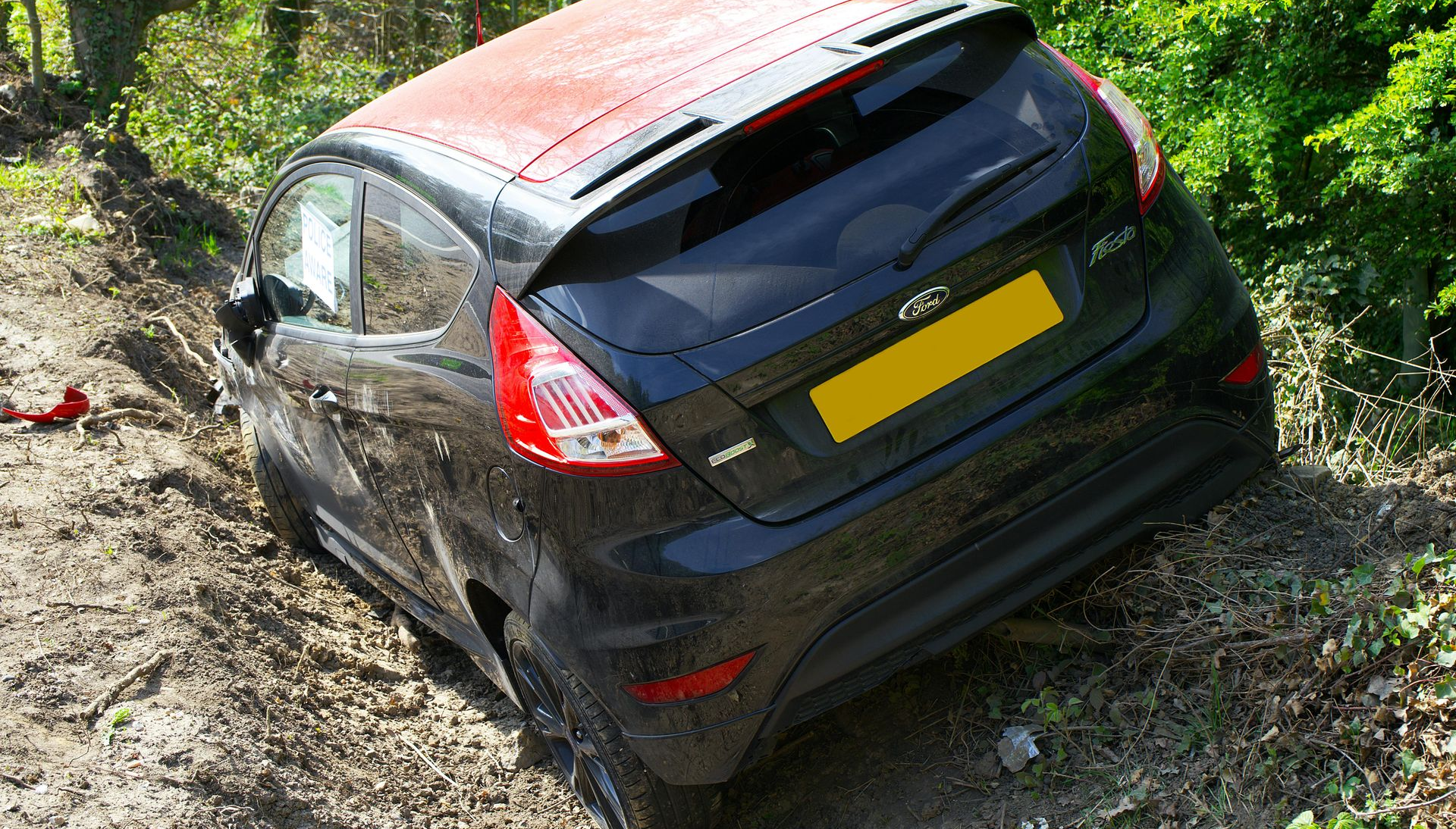 A dark Ford Fiesta car partially submerged in a muddy ditch alongside a wooded area.