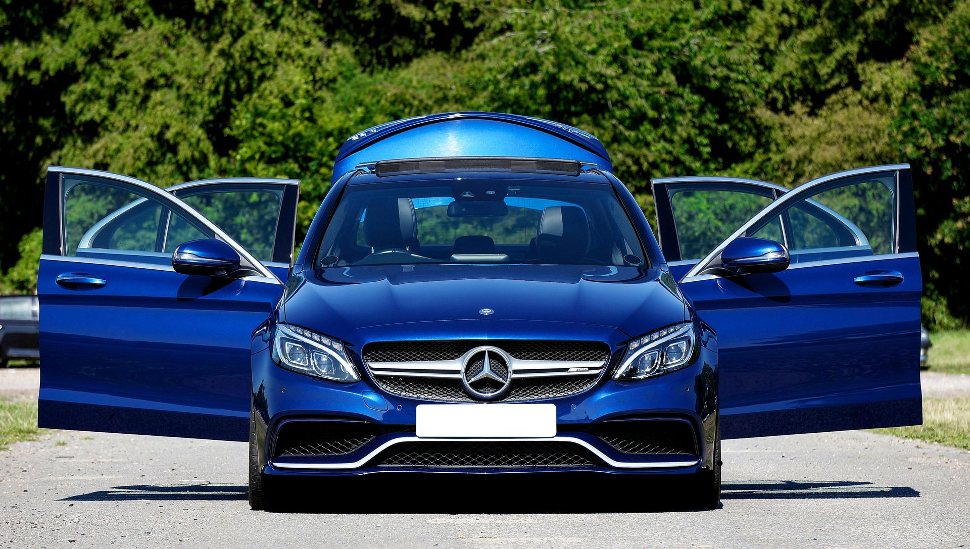 Blue Mercedes sedan with both front doors open on a road, trees in the background