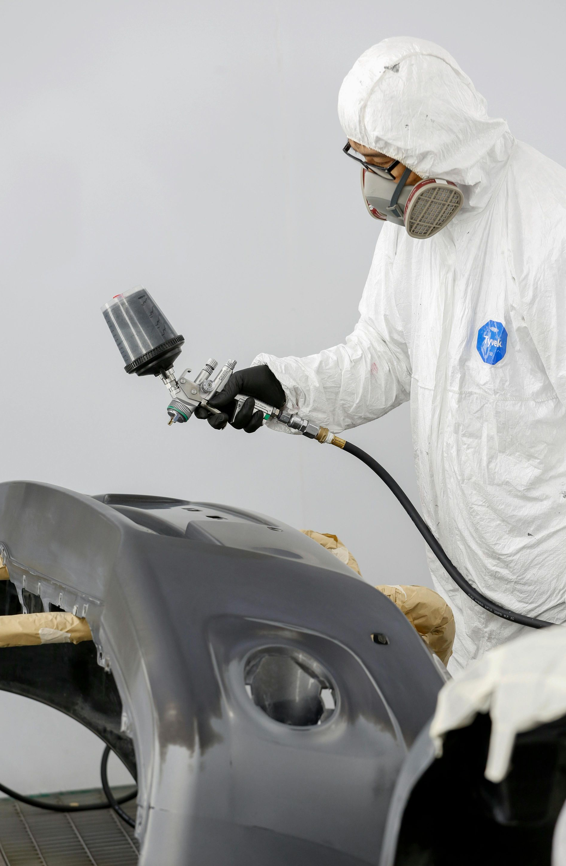 A person in protective coveralls and a respirator uses a spray gun to paint a gray vehicle bumper.