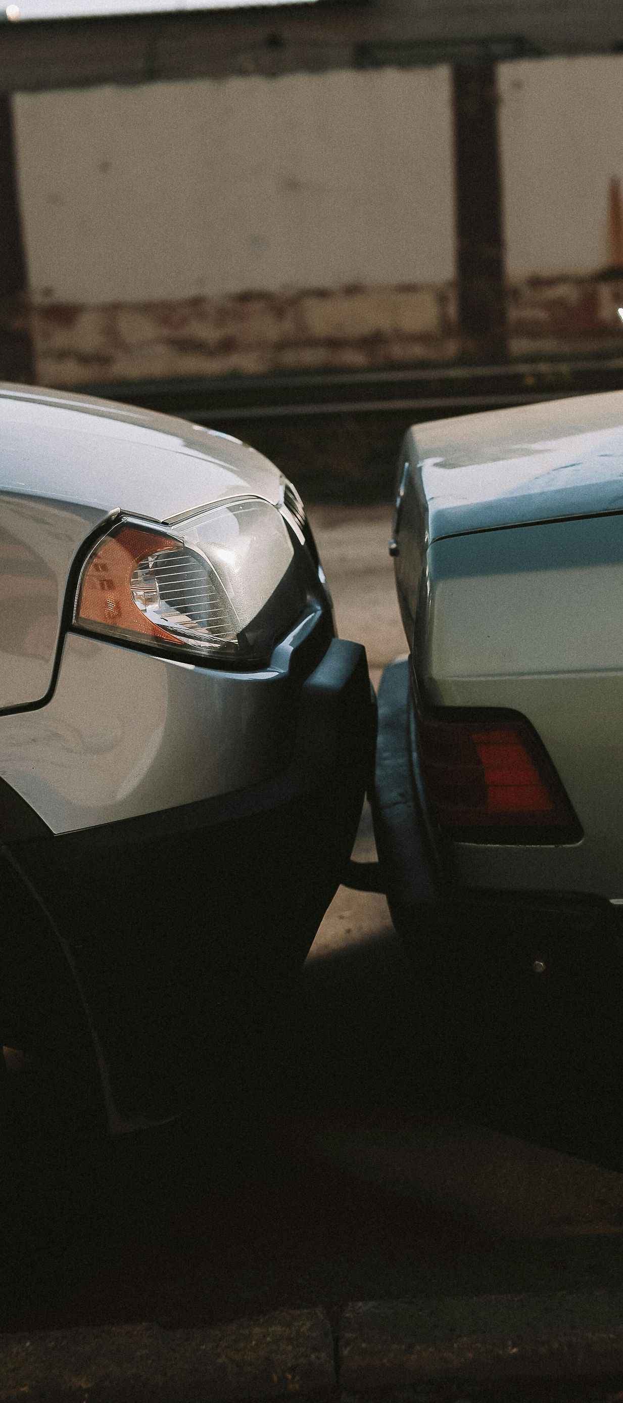 Two cars, one silver, one green, bumper to bumper. Brick wall in the background.